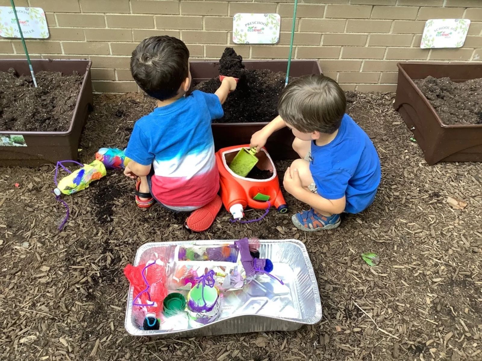 Two SELA children kneel at raised garden beds, scooping soil into pots during an outdoor planting activity.