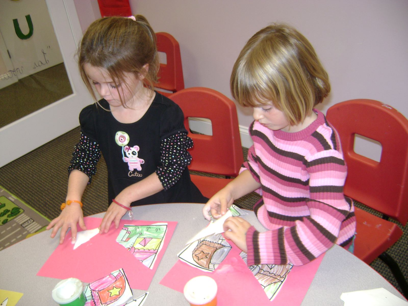 Two young SELA students in art smocks paint together at a classroom table, with Spanish color words posted on the wall behind them.