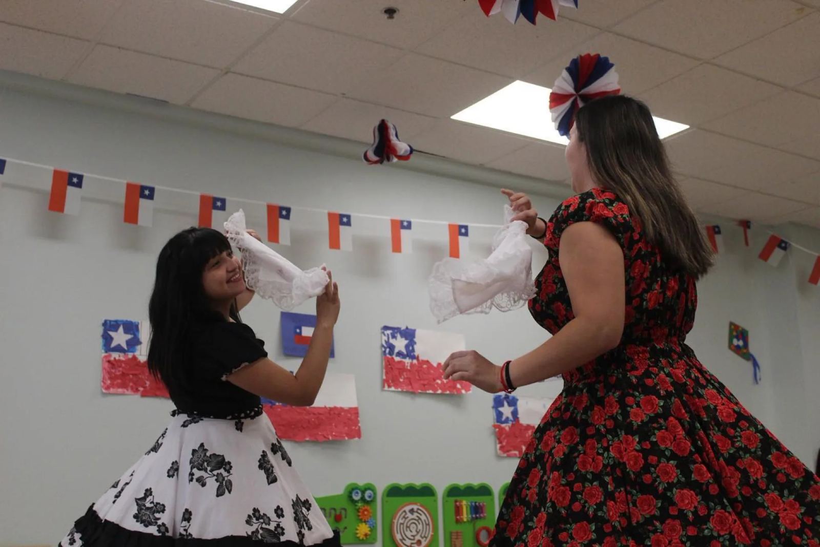 A SELA student and a teacher in a rose-print dress dance the Chilean cueca, waving white handkerchiefs beneath strings of Chilean flags.