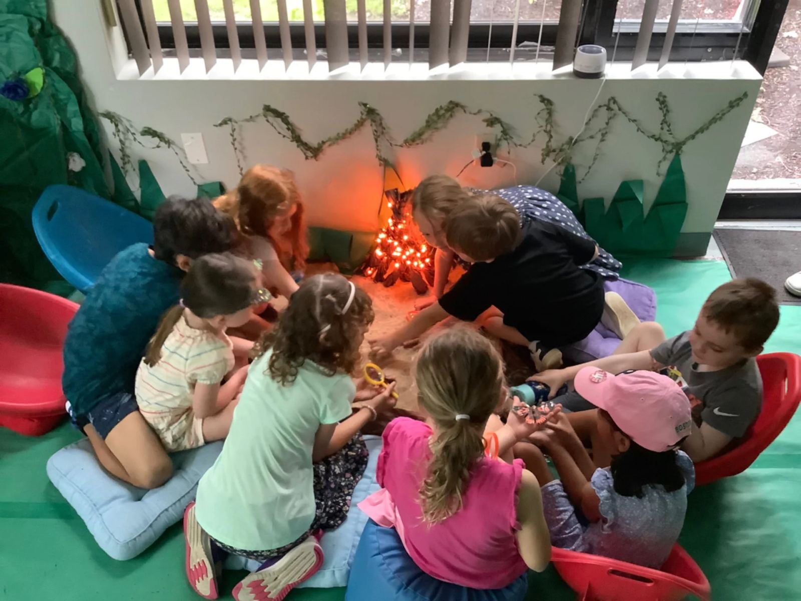 SELA children gather close around a pretend indoor campfire of lights and logs during a summer story circle.