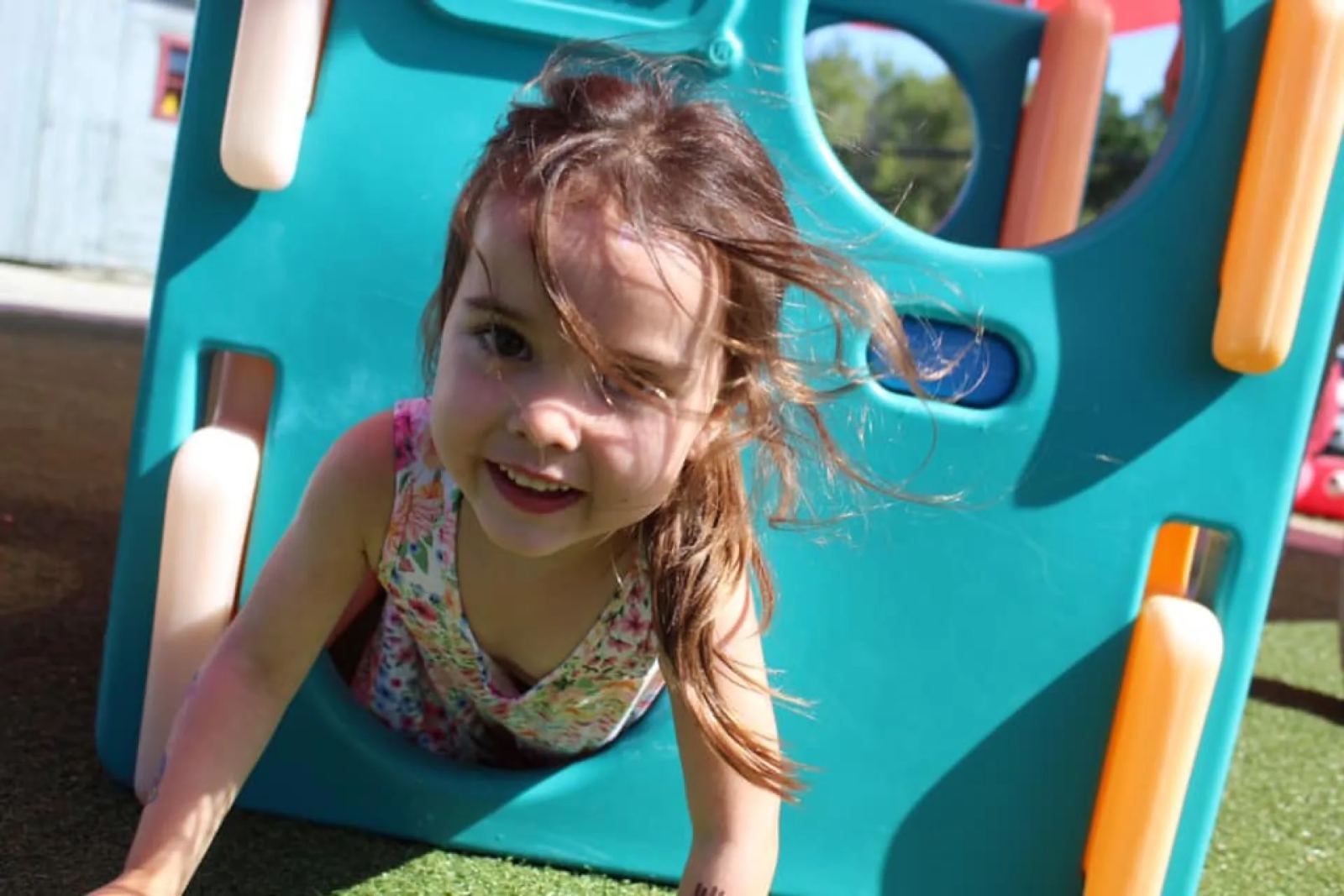 A SELA child grins as she crawls out of a teal playground tunnel, hair caught mid-motion in the wind.