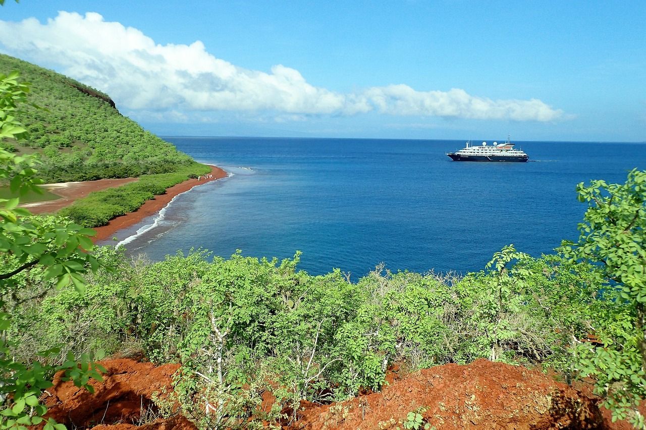 Green hillside and red-sand beach on Rábida Island in the Galápagos, with a ship offshore