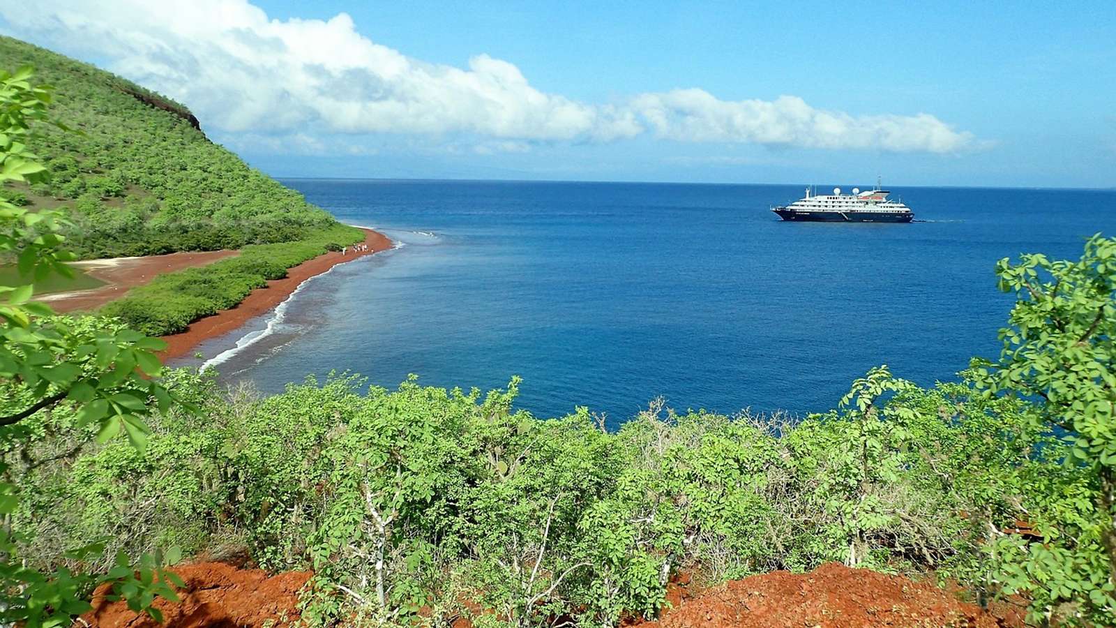 Green hillside and red-sand beach on Rábida Island in the Galápagos, with a ship offshore