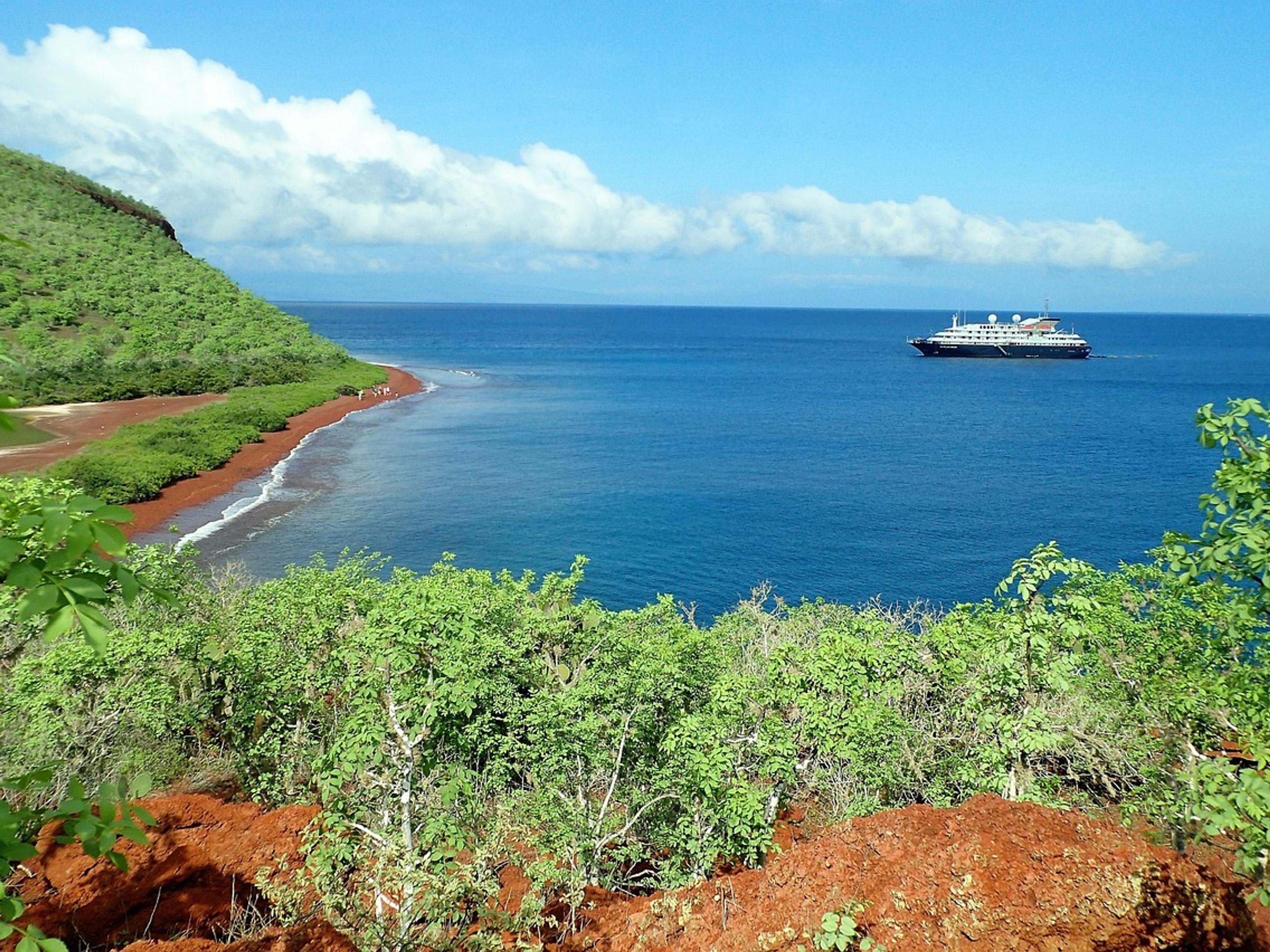 Green hillside and red-sand beach on Rábida Island in the Galápagos, with a ship offshore