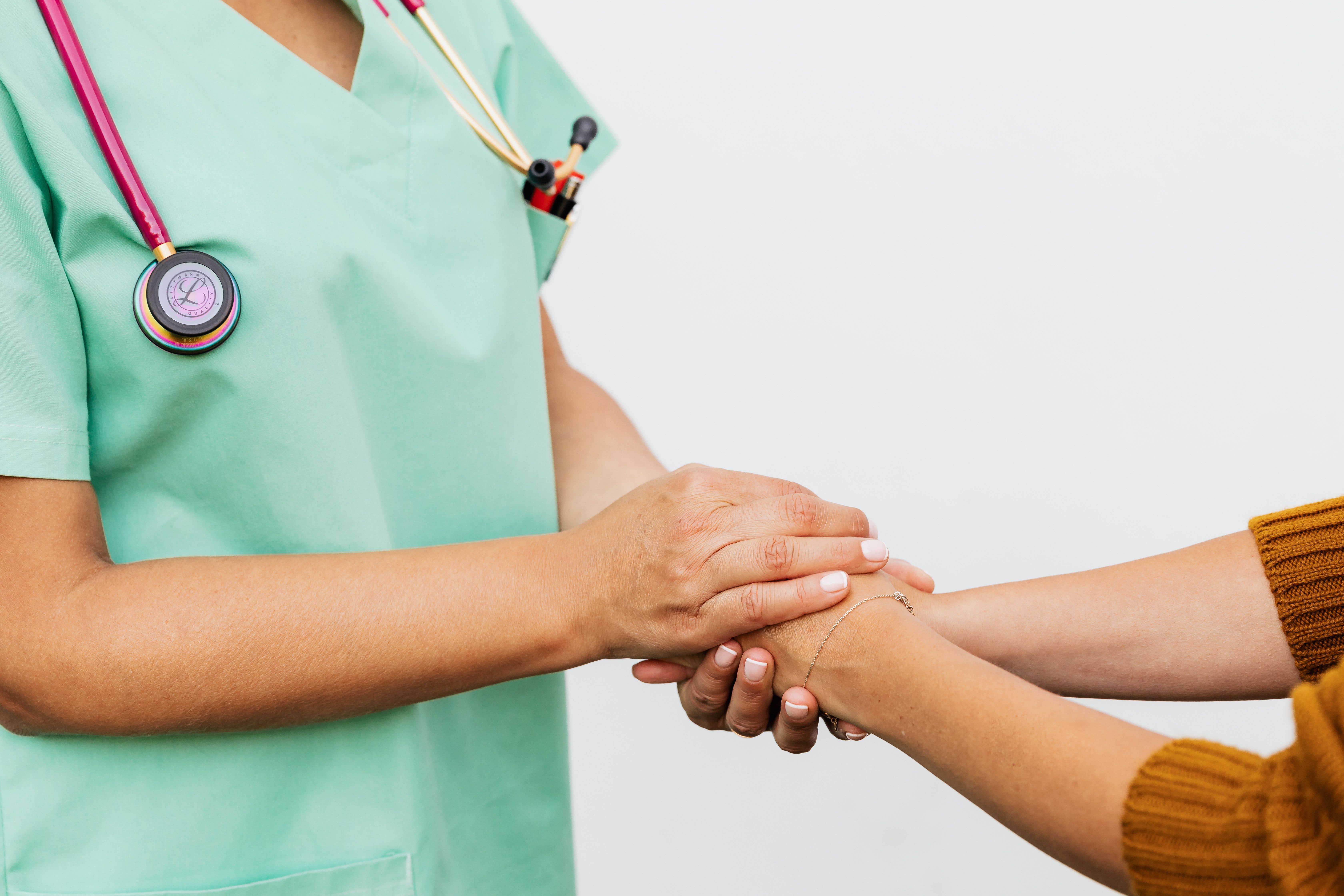 A nurse in green scrubs gently holding a patient's hand and wrist in a comforting gesture.