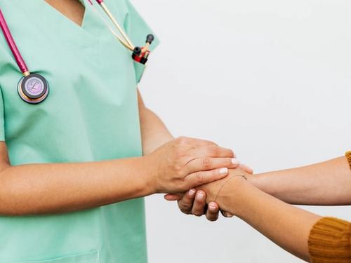 A nurse in green scrubs gently holding a patient's hand and wrist in a comforting gesture.