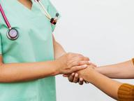 A nurse in green scrubs gently holding a patient's hand and wrist in a comforting gesture.