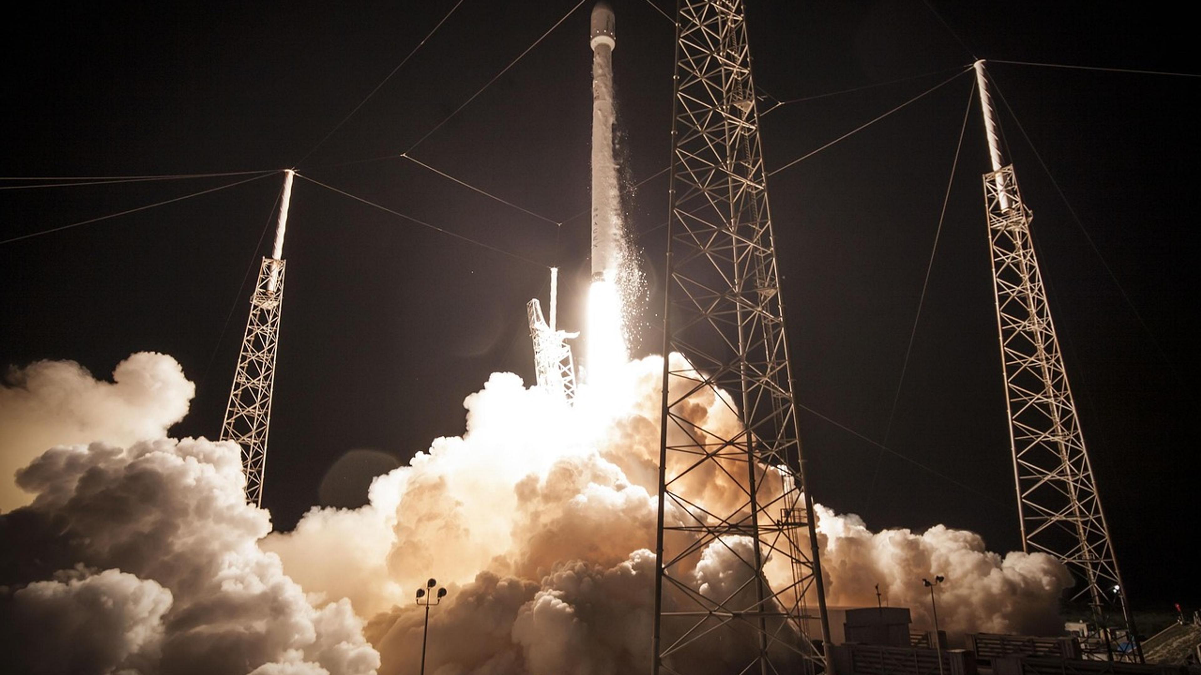 A rocket lifting off at night in a cloud of exhaust beside its launch towers