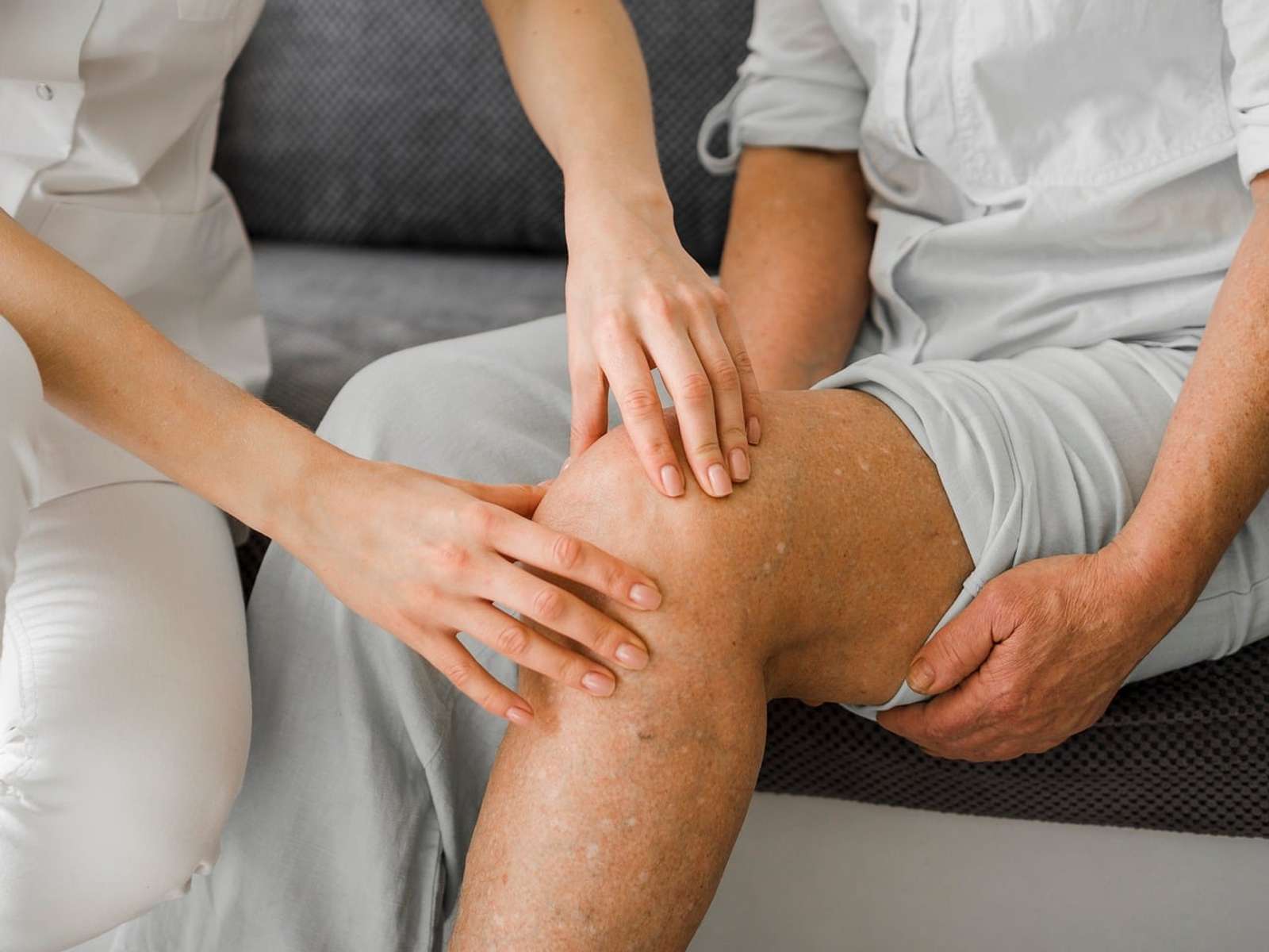 A physical therapist's hands examining the knee of an older patient seated on a couch