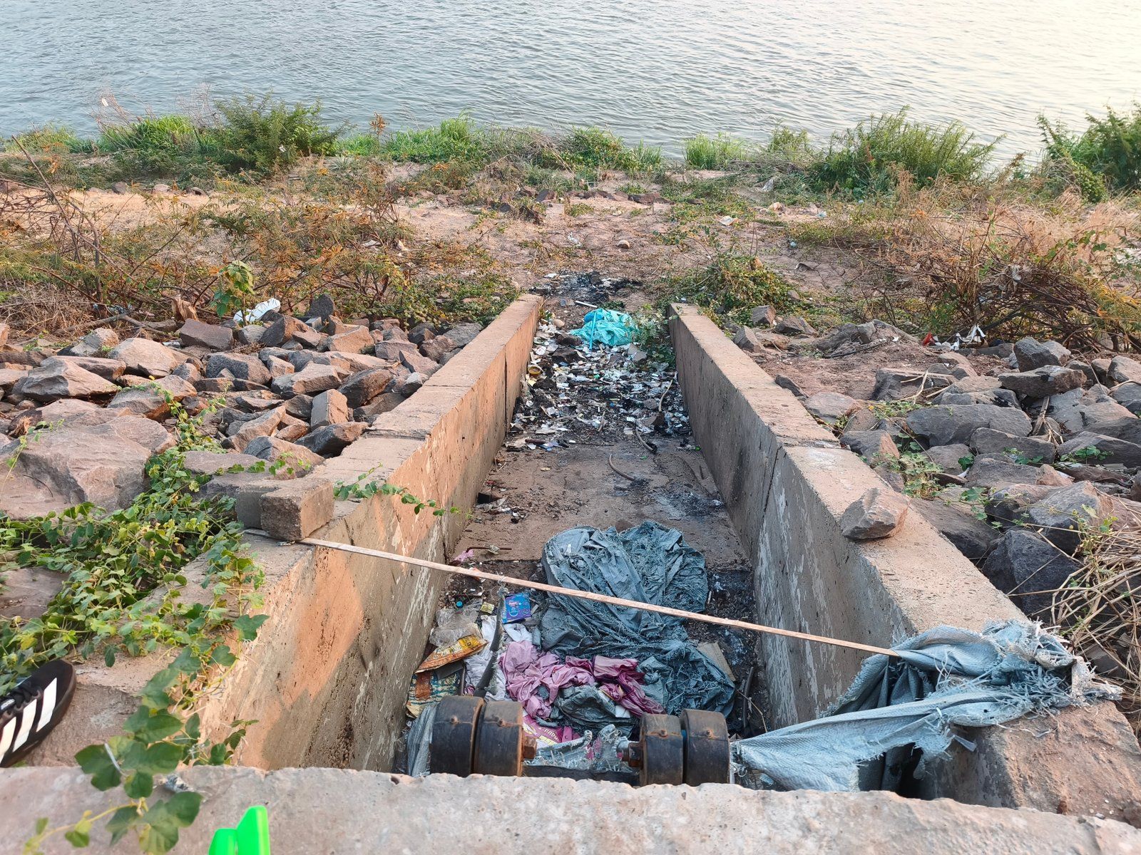 Discarded plastic bags and litter in a concrete drainage channel on a rocky riverbank