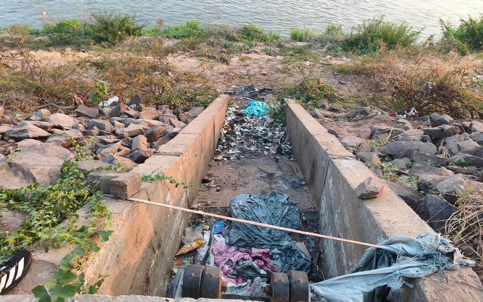 Discarded plastic bags and litter in a concrete drainage channel on a rocky riverbank