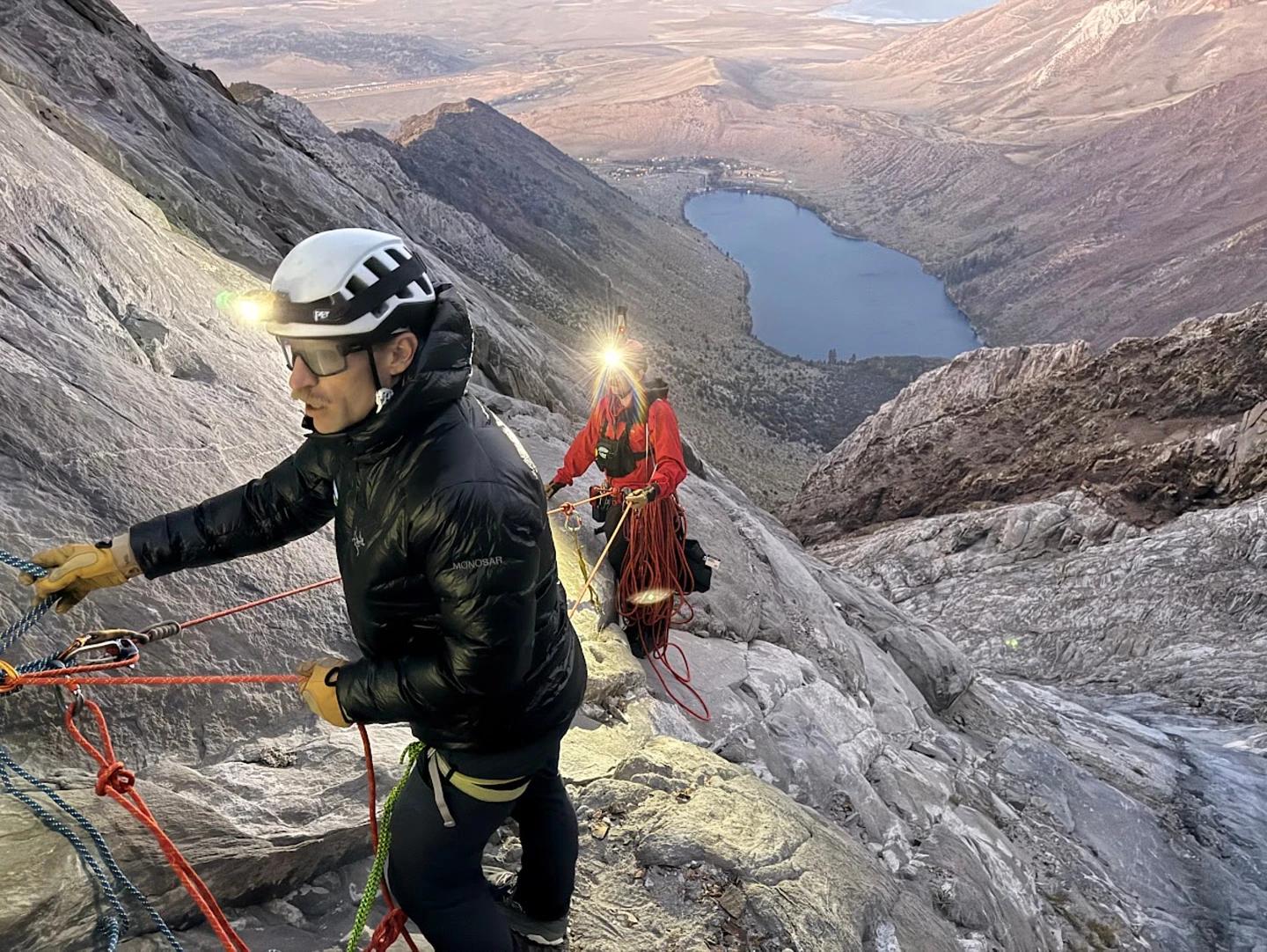 A rescuer in high-visibility orange gear rappels down a rugged rocky mountain slope