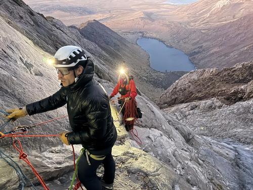A rescuer in high-visibility orange gear rappels down a rugged rocky mountain slope