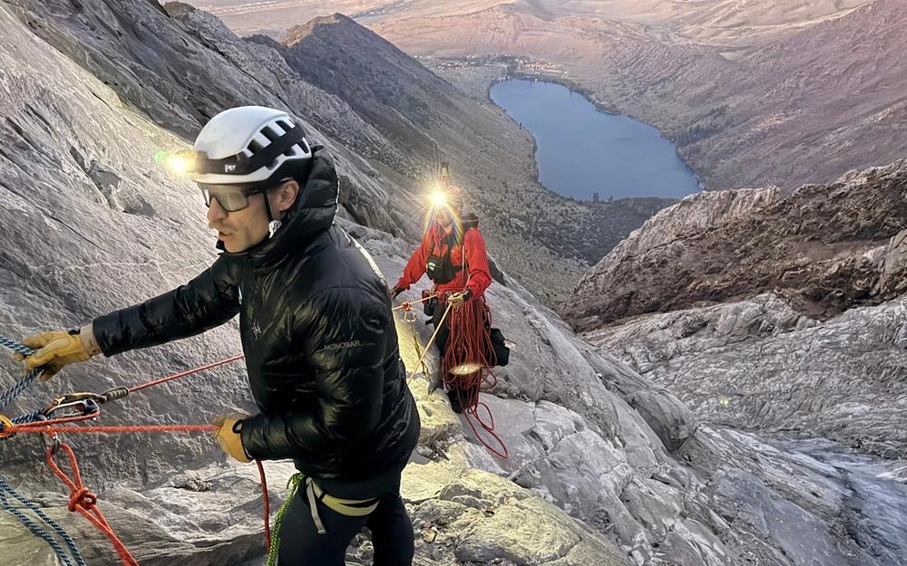 A rescuer in high-visibility orange gear rappels down a rugged rocky mountain slope