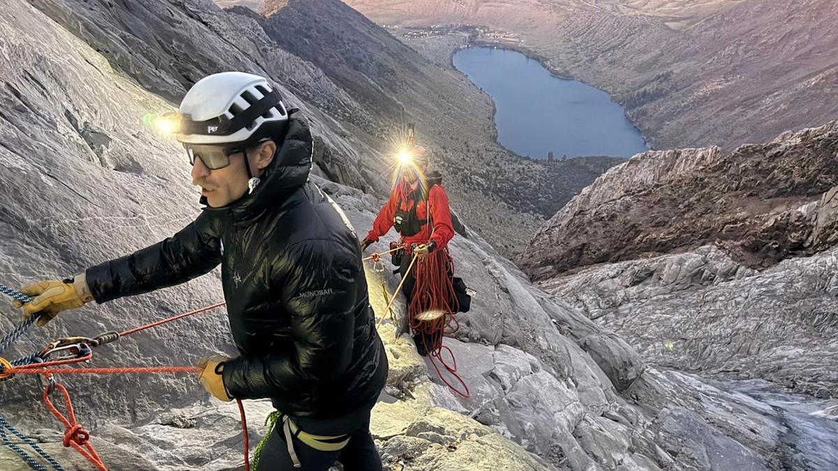 A rescuer in high-visibility orange gear rappels down a rugged rocky mountain slope