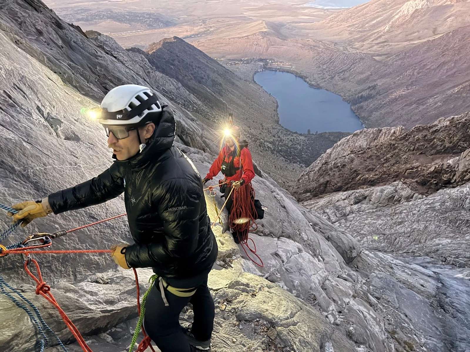 A rescuer in high-visibility orange gear rappels down a rugged rocky mountain slope