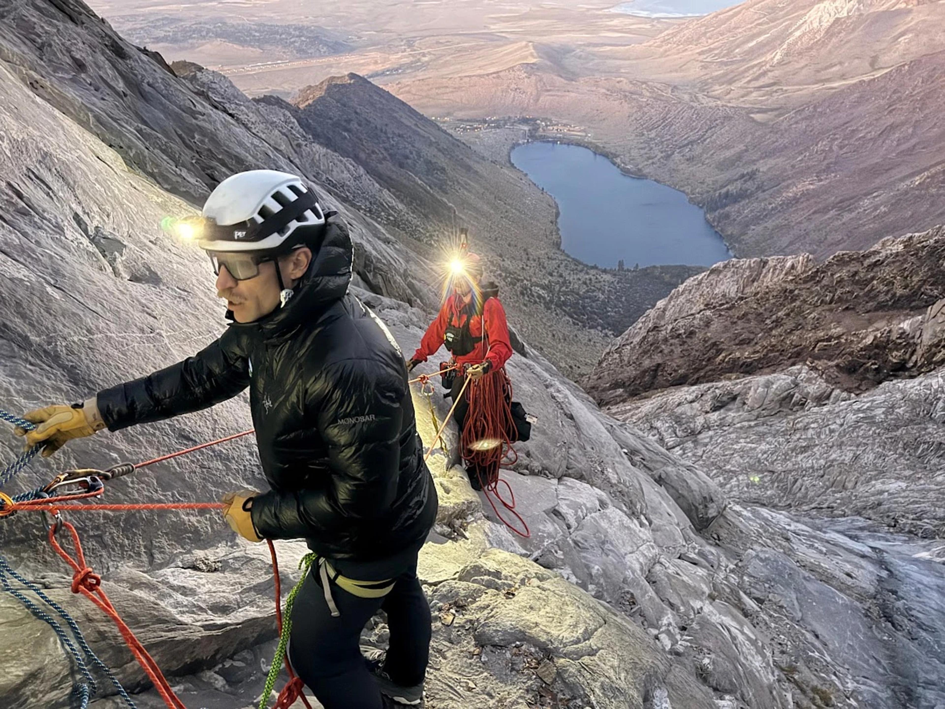 A rescuer in high-visibility orange gear rappels down a rugged rocky mountain slope