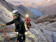 A rescuer in high-visibility orange gear rappels down a rugged rocky mountain slope