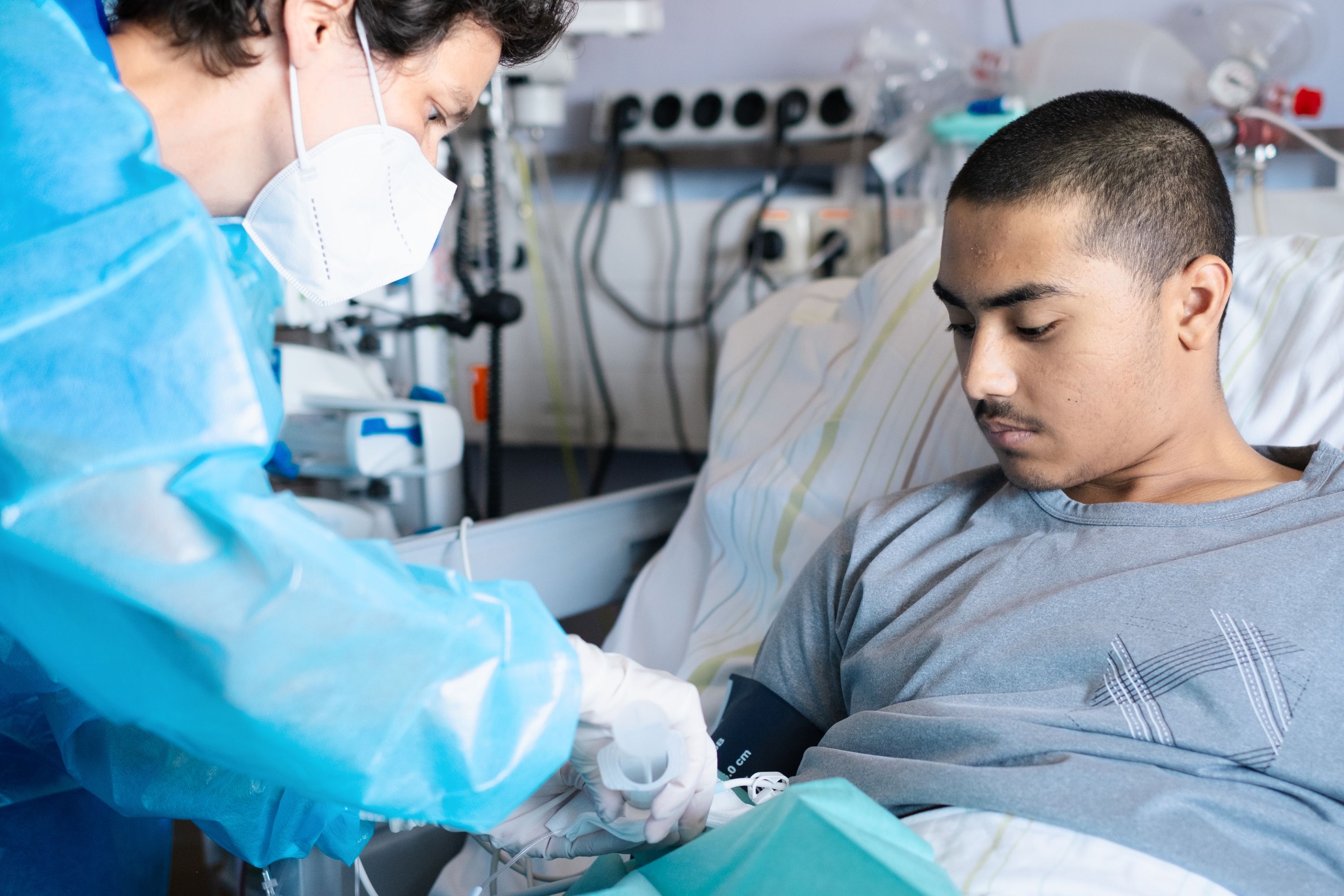 A doctor in a blue protective gown and mask connects an infusion line to a young man lying in a hospital bed