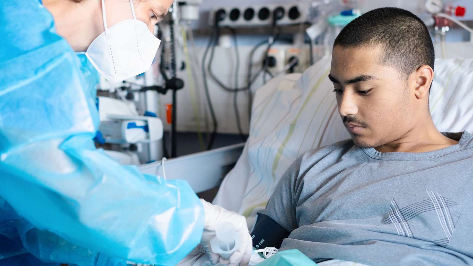 A doctor in a blue protective gown and mask connects an infusion line to a young man lying in a hospital bed