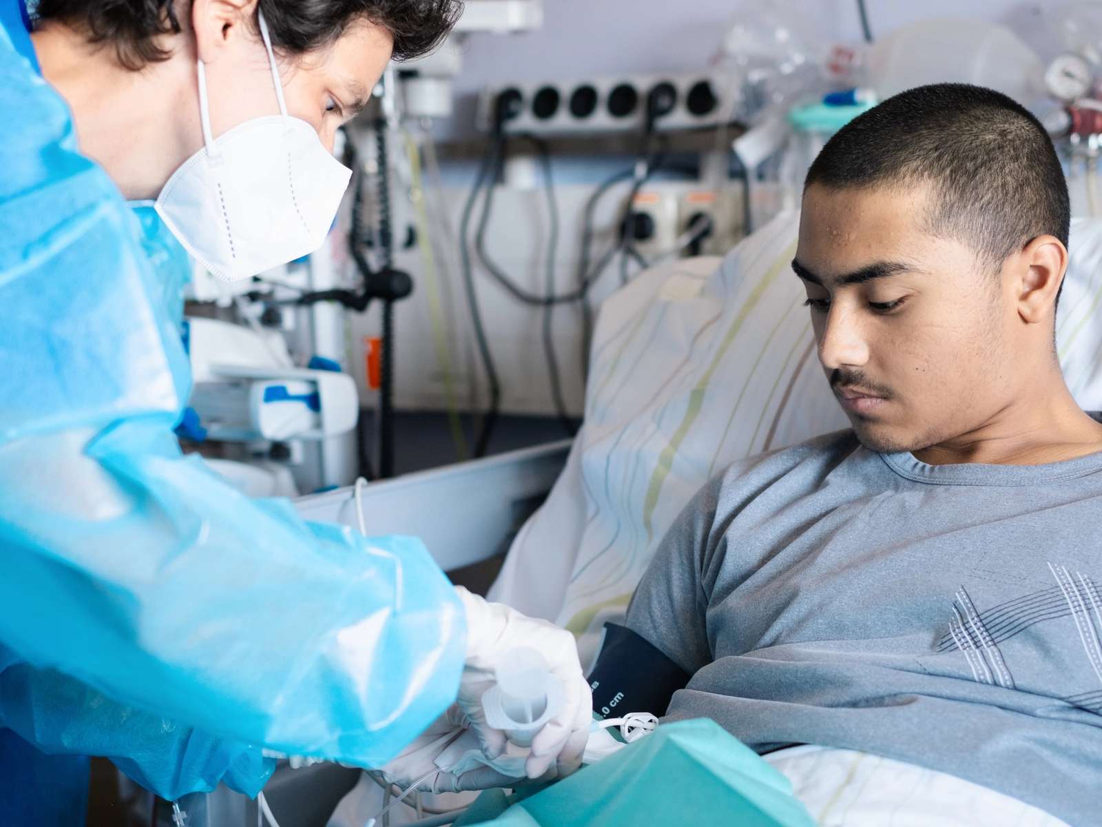 A doctor in a blue protective gown and mask connects an infusion line to a young man lying in a hospital bed