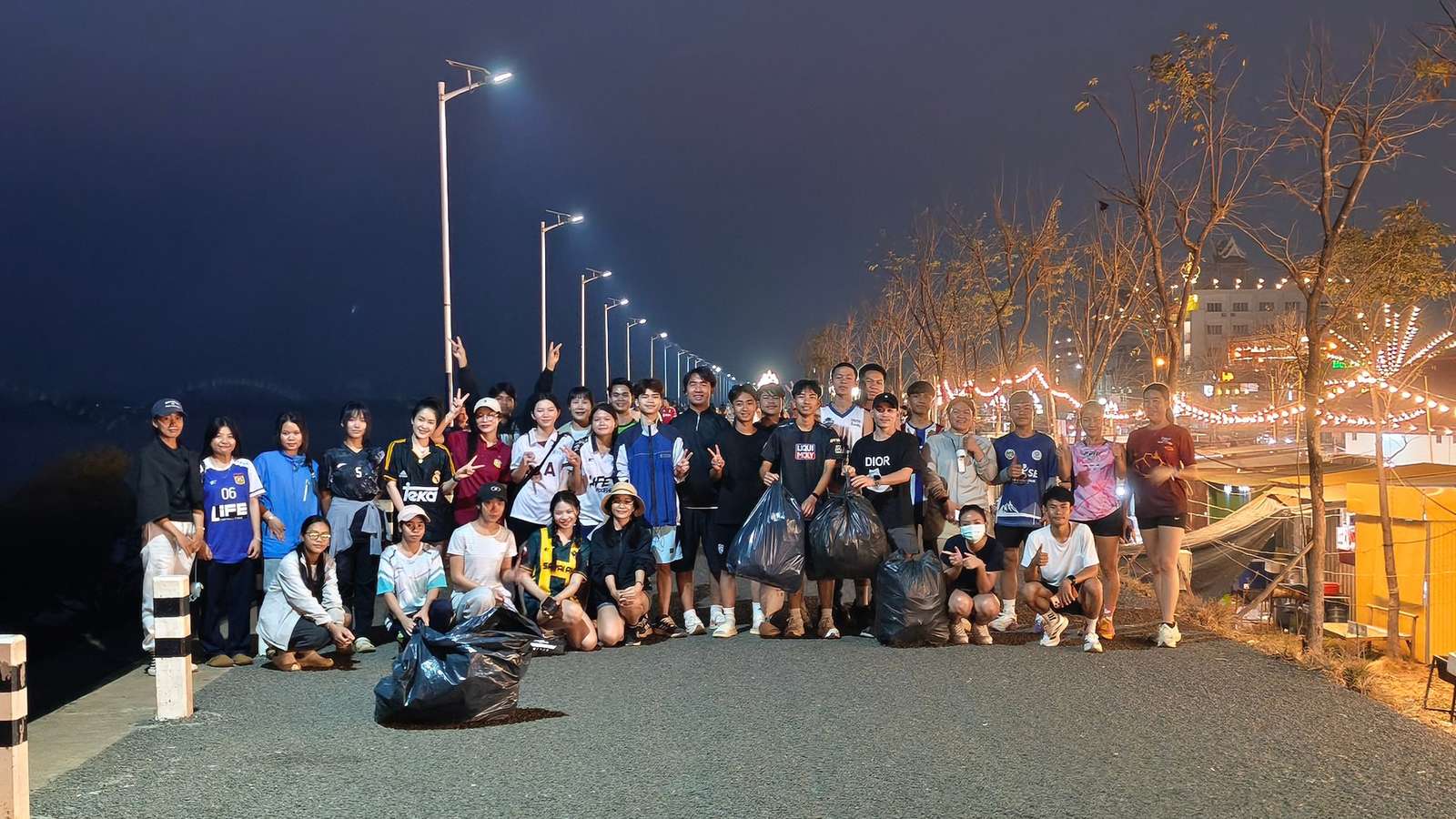 A large group of volunteers with filled black trash bags posing on a lit riverside promenade at dusk