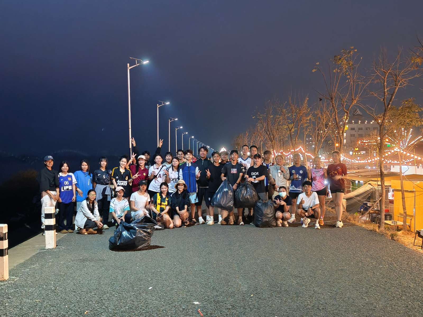 A large group of volunteers with filled black trash bags posing on a lit riverside promenade at dusk