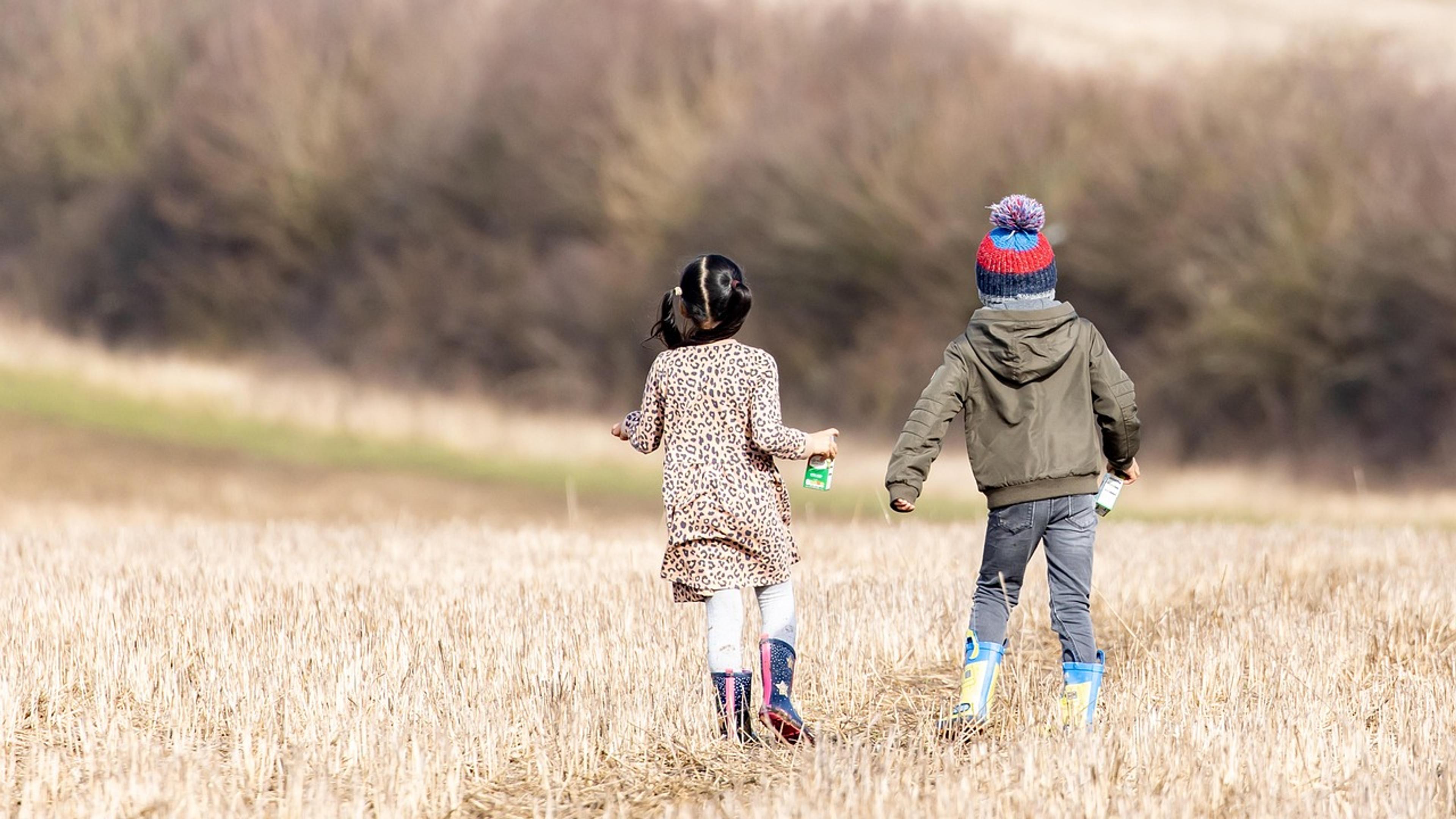 Two young children in boots and jackets running across a mown field