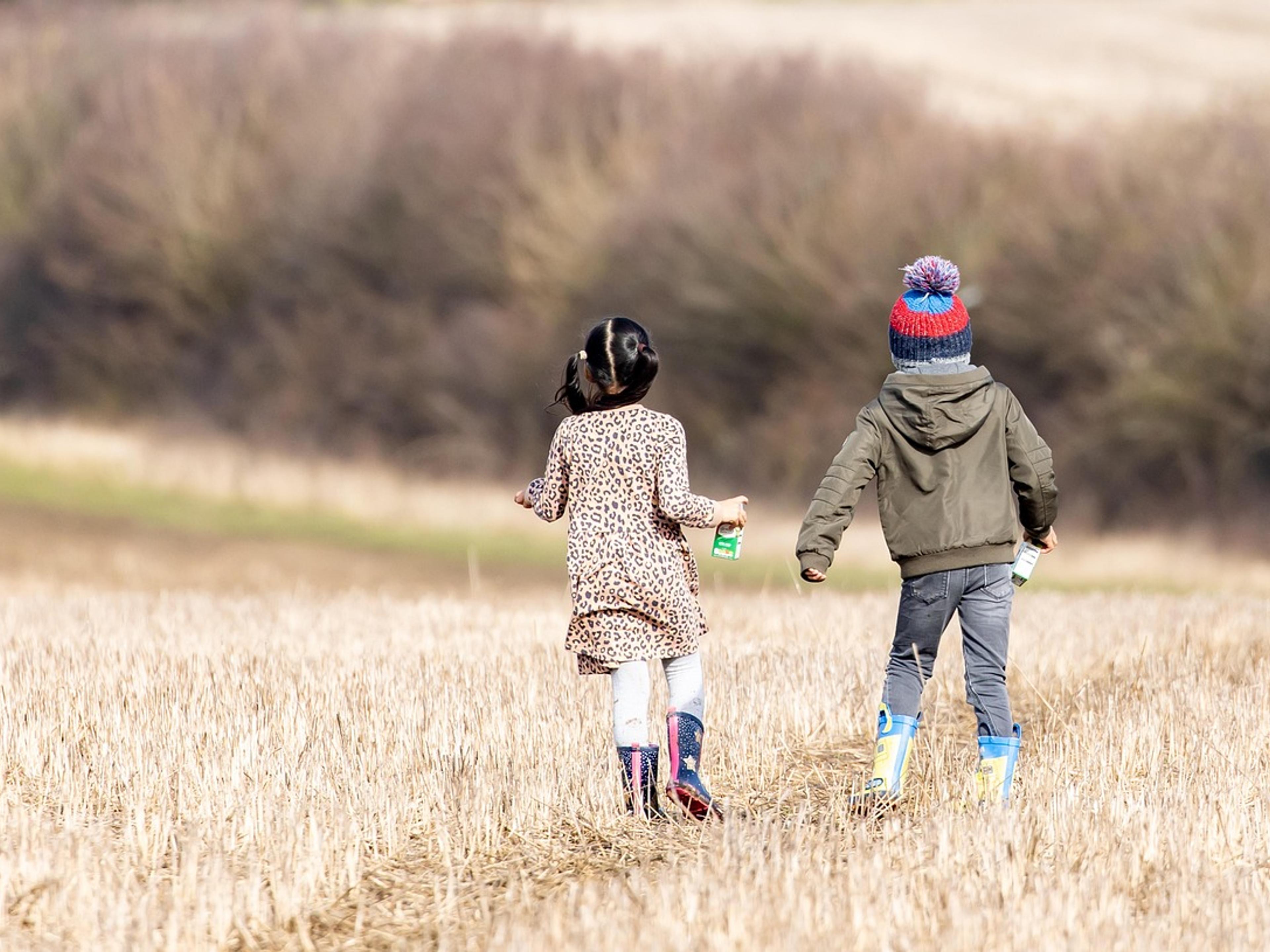 Two young children in boots and jackets running across a mown field