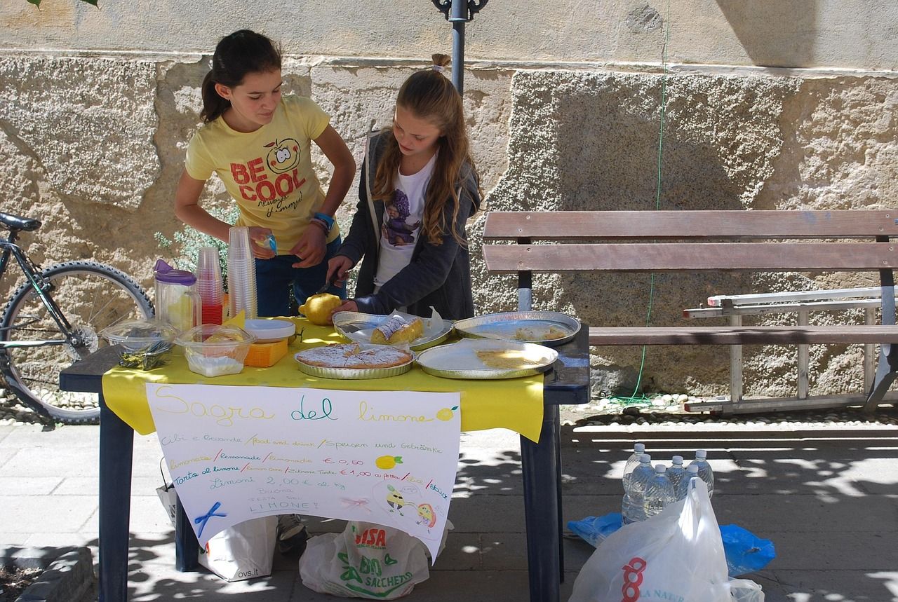 Two girls run a homemade lemonade stand at a table with a hand-drawn sign on a sunny street