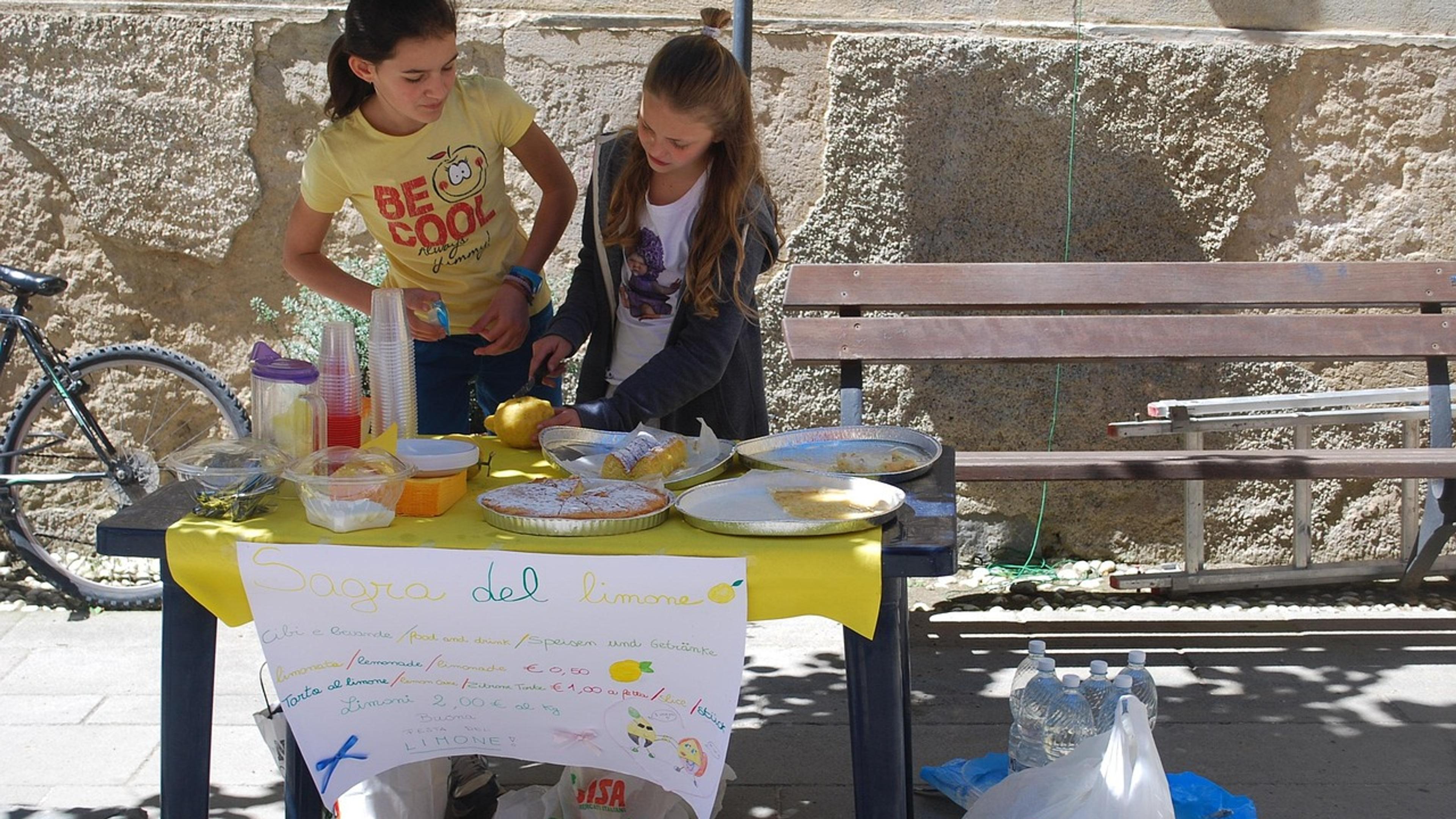 Two girls run a homemade lemonade stand at a table with a hand-drawn sign on a sunny street