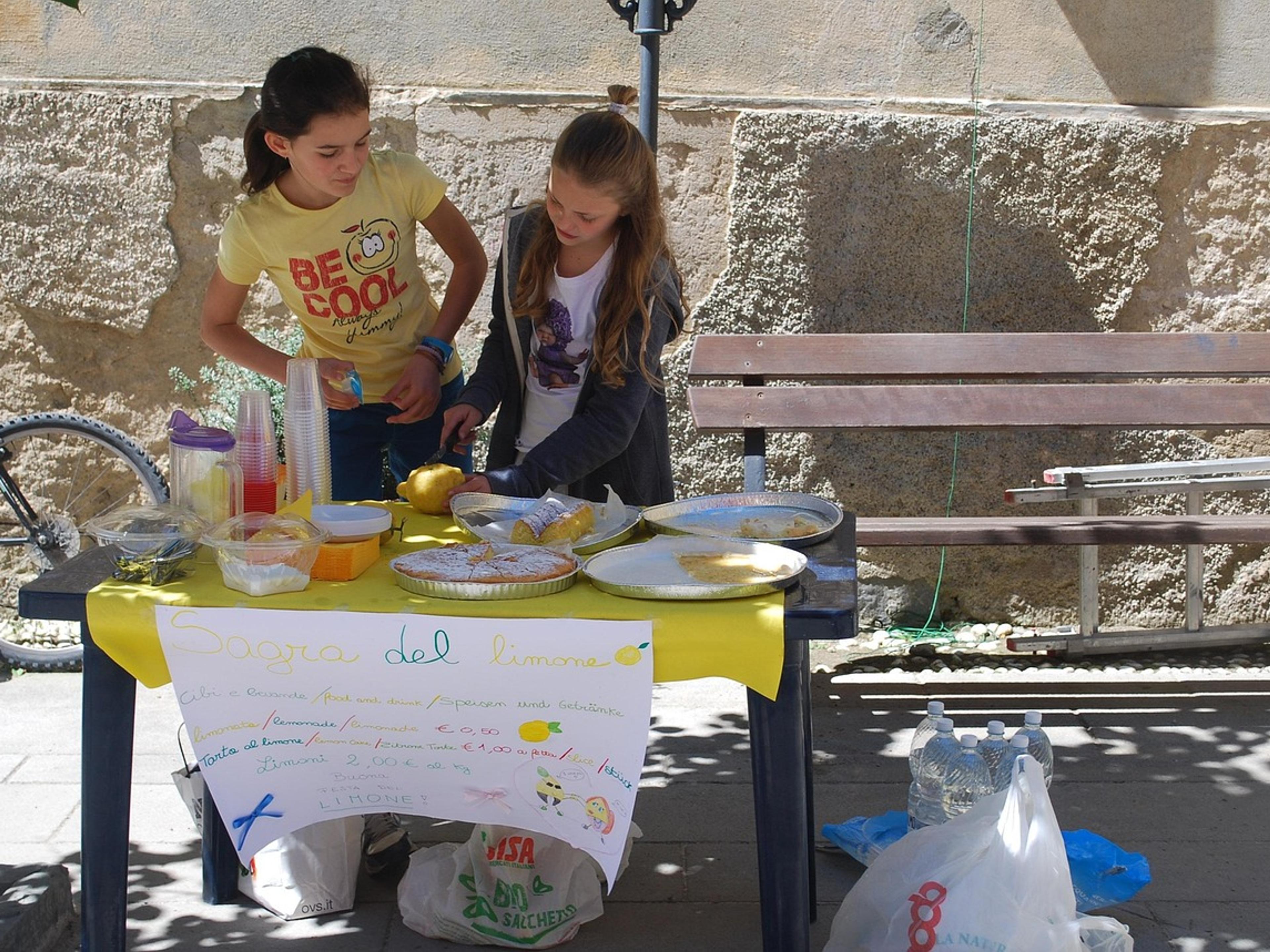 Two girls run a homemade lemonade stand at a table with a hand-drawn sign on a sunny street