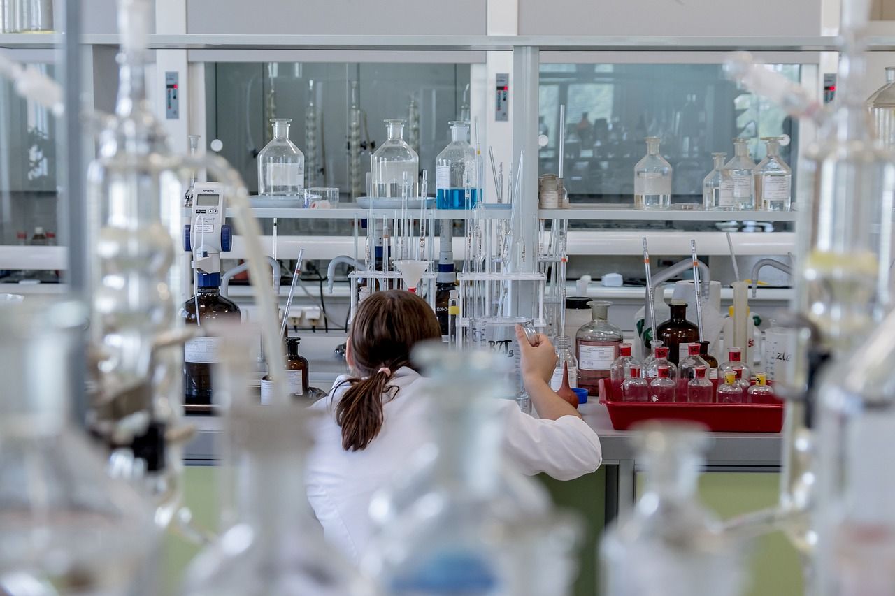 A researcher in a white lab coat works at a bench in a chemistry laboratory, seen past rows of glass bottles
