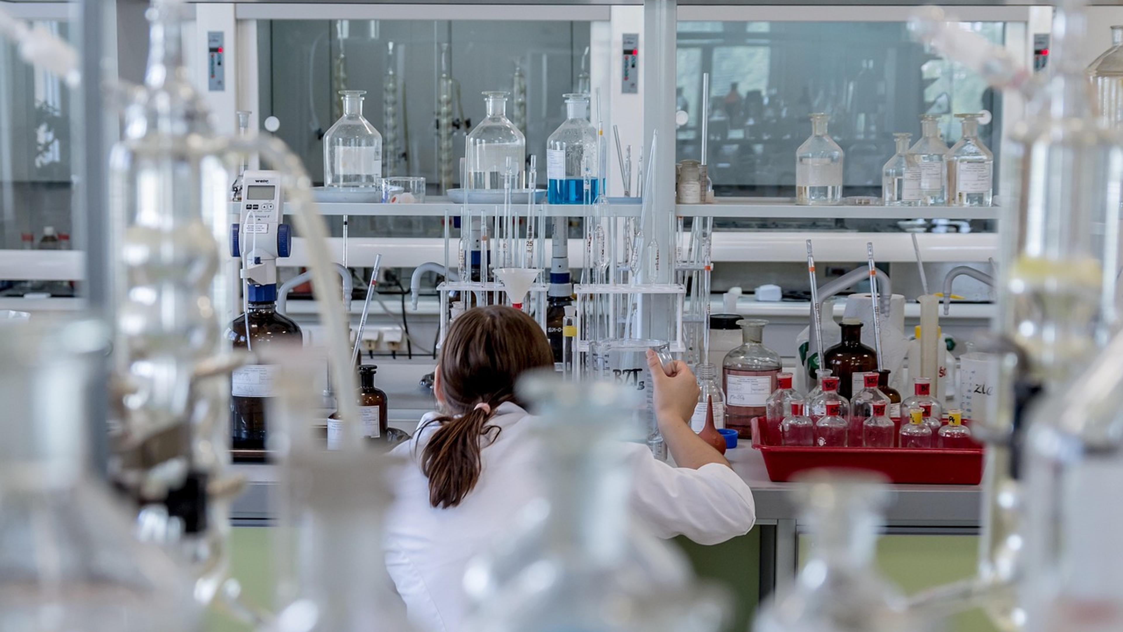 A researcher in a white lab coat works at a bench in a chemistry laboratory, seen past rows of glass bottles
