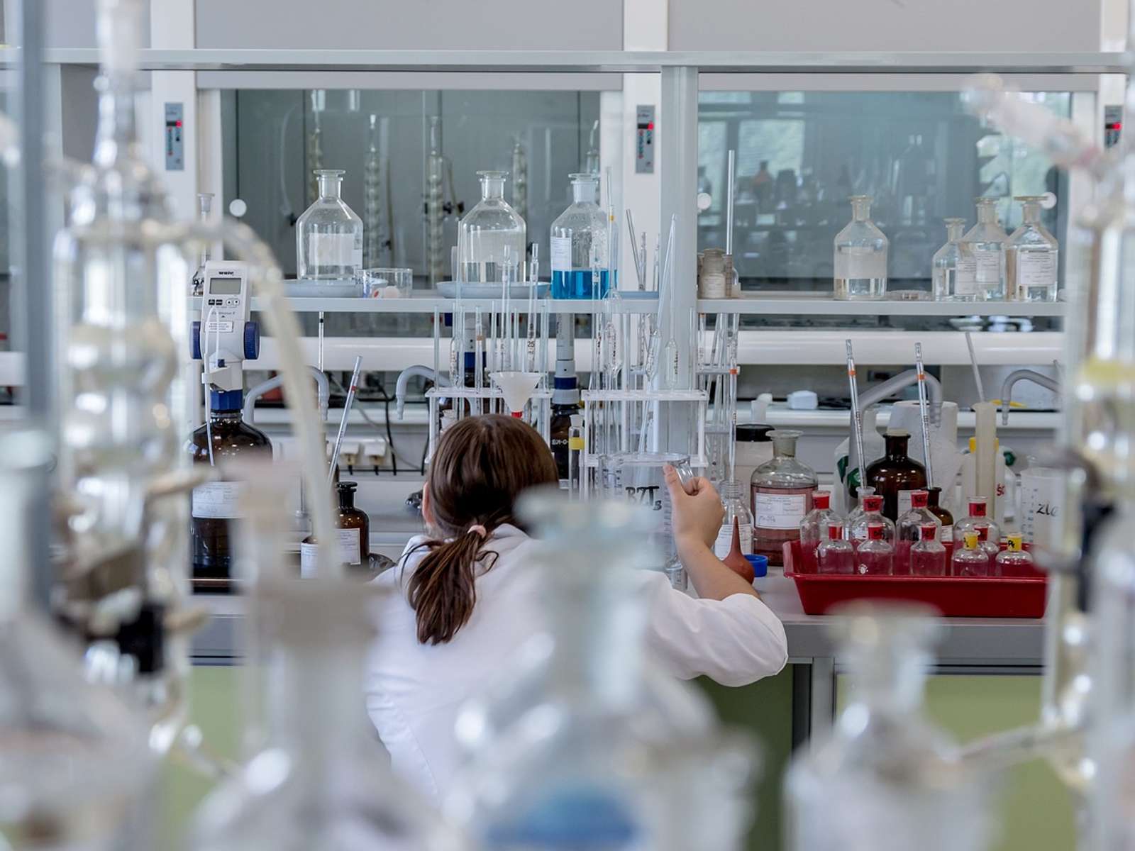 A researcher in a white lab coat works at a bench in a chemistry laboratory, seen past rows of glass bottles