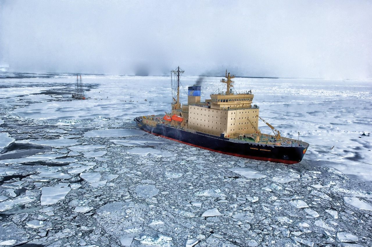 An icebreaker ship pushing through broken sea ice, with a sailing ship in the distance