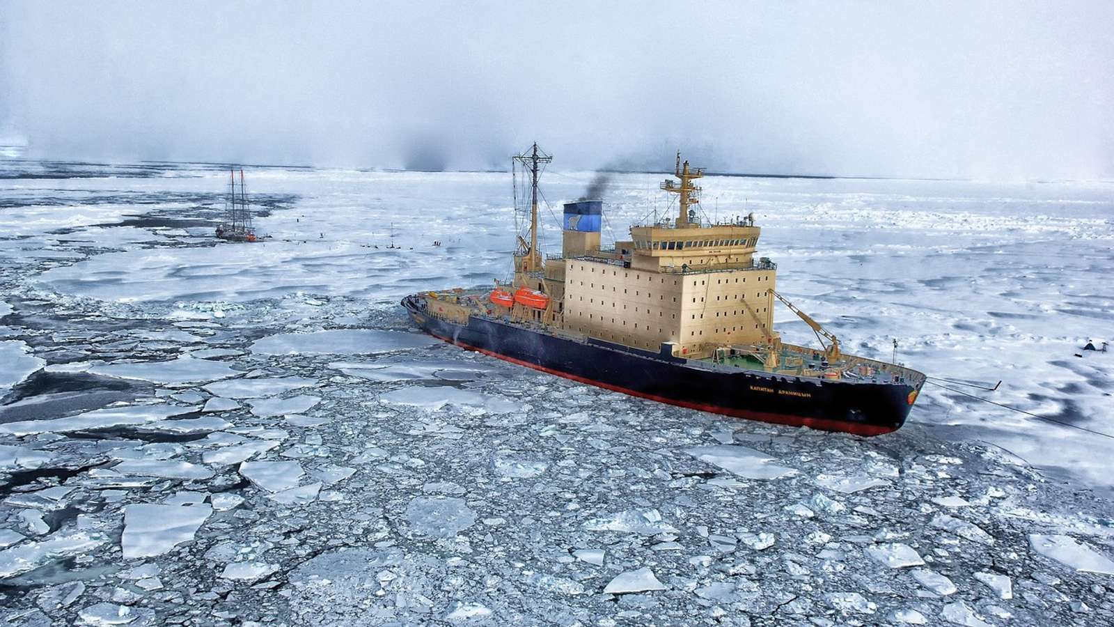 An icebreaker ship pushing through broken sea ice, with a sailing ship in the distance