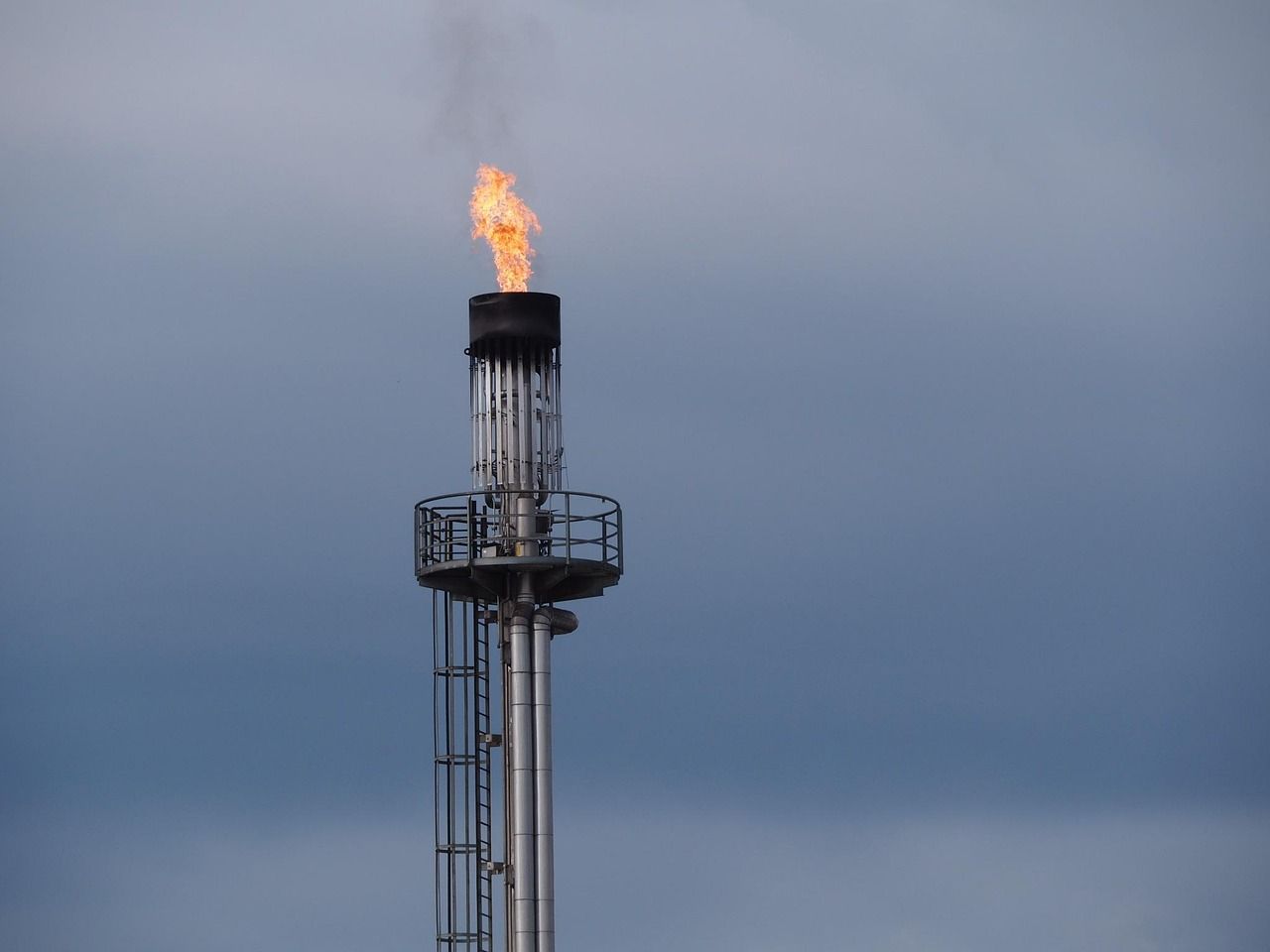 A gas flare burning at the top of a tall industrial stack against a grey sky