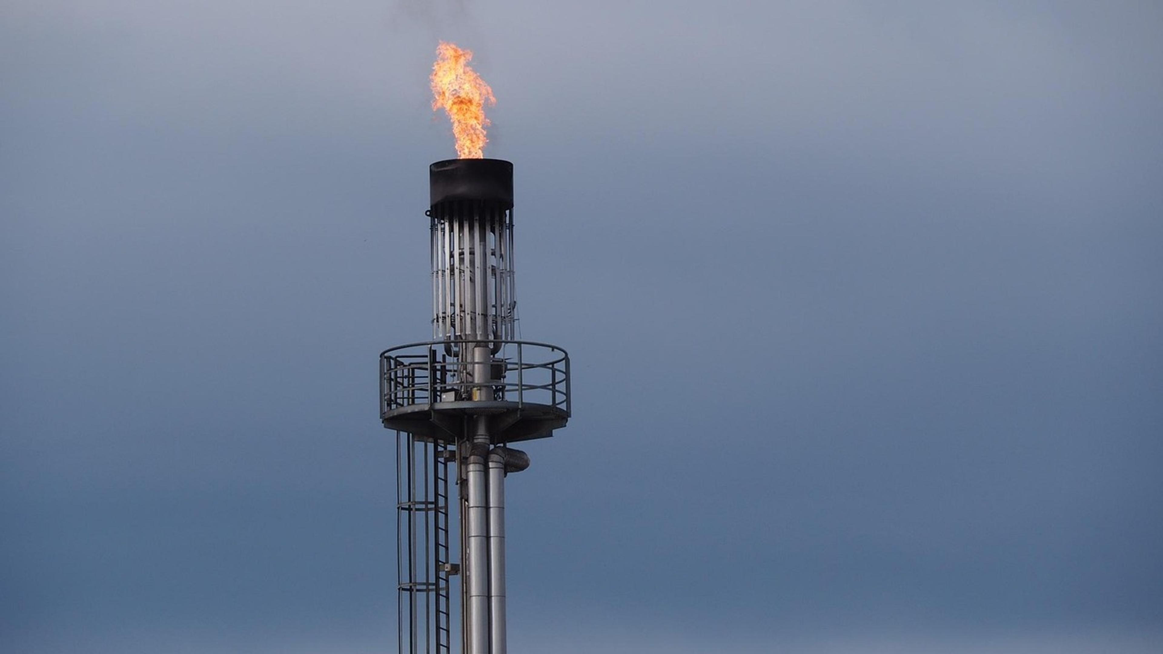 A gas flare burning at the top of a tall industrial stack against a grey sky