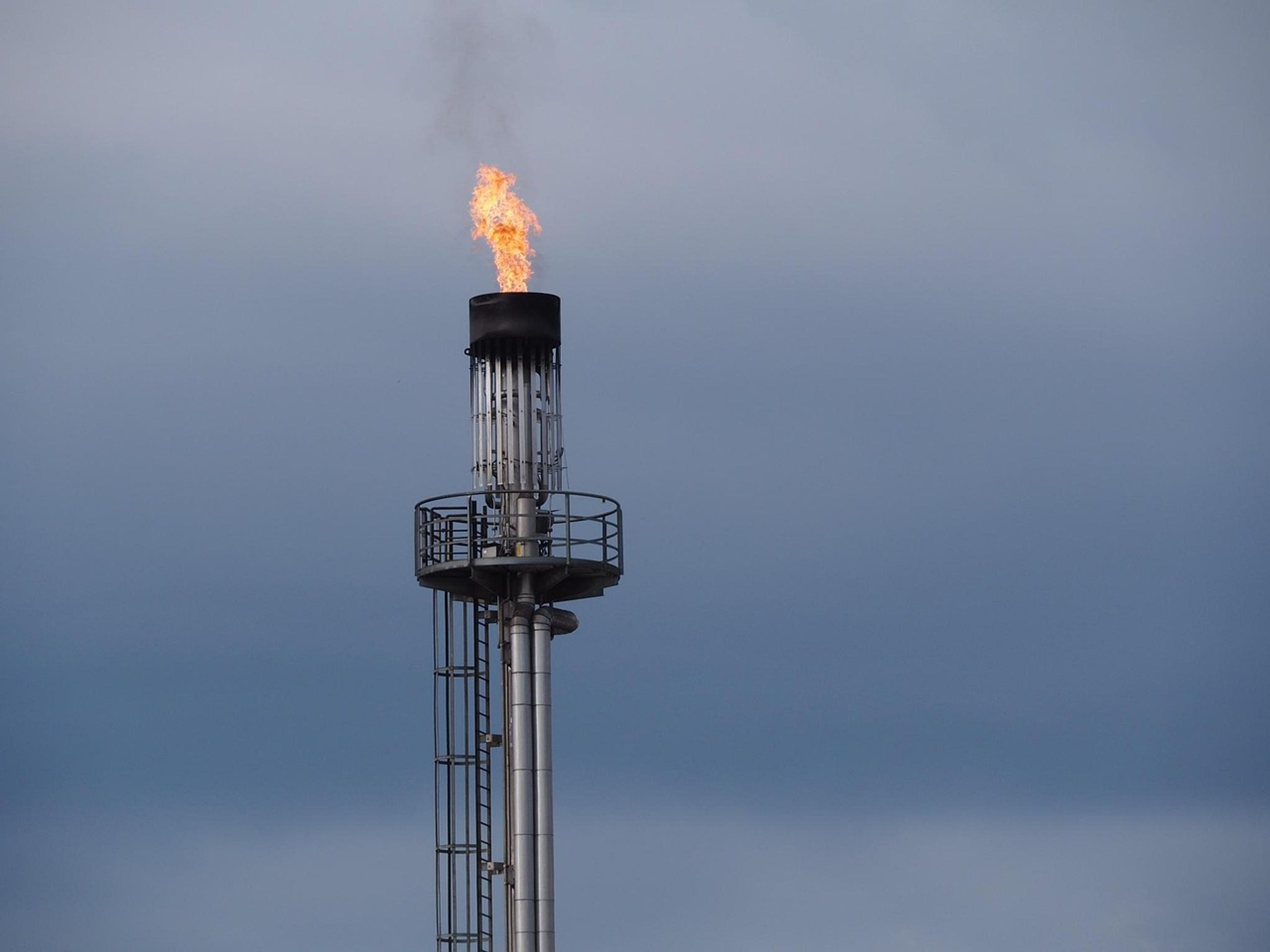 A gas flare burning at the top of a tall industrial stack against a grey sky