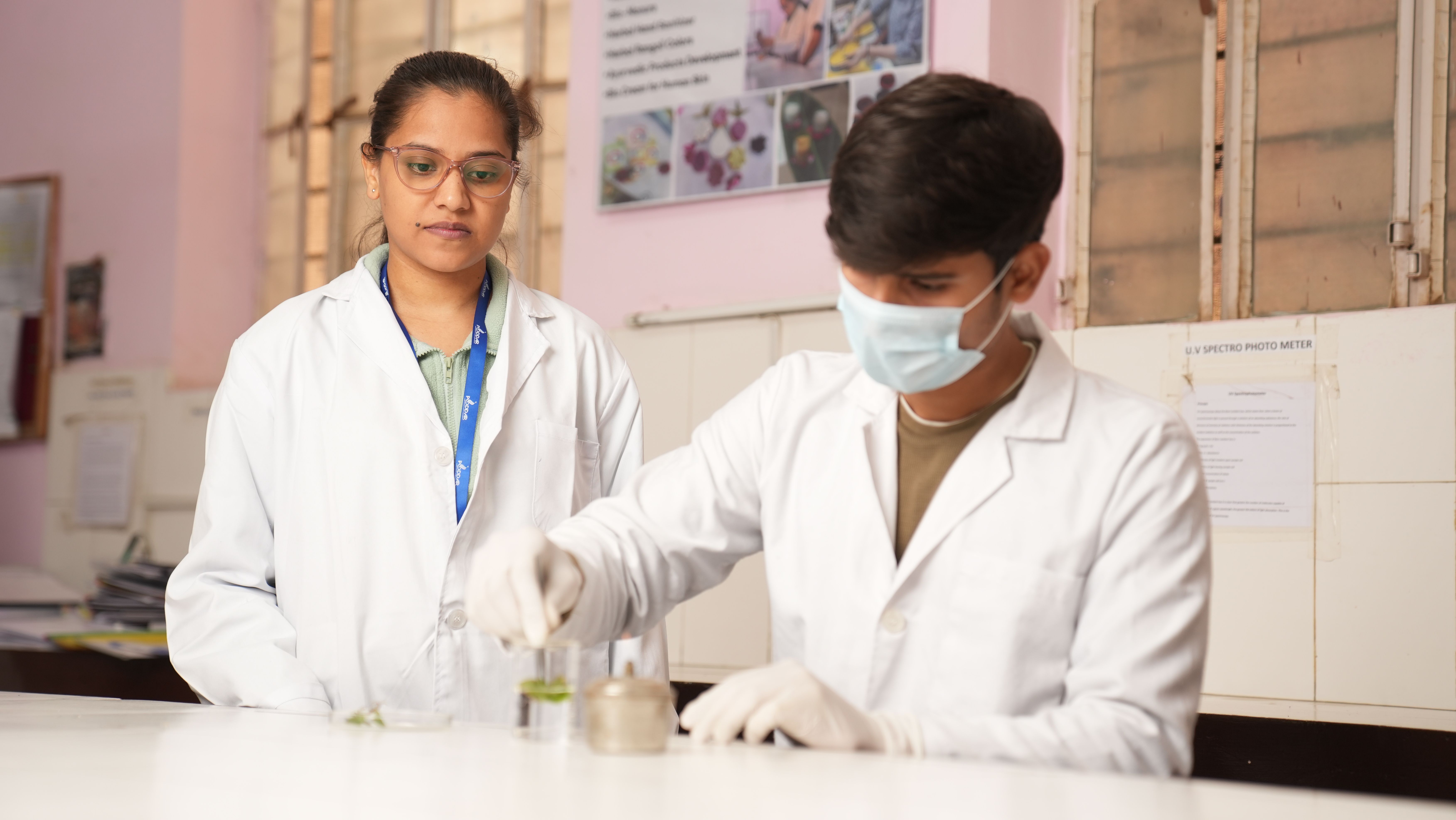 Two scientists in white lab coats and safety gloves working together at a laboratory bench with glassware and research equipment.
