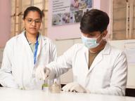 Two scientists in white lab coats and safety gloves working together at a laboratory bench with glassware and research equipment.