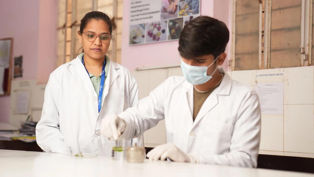 Two scientists in white lab coats and safety gloves working together at a laboratory bench with glassware and research equipment.