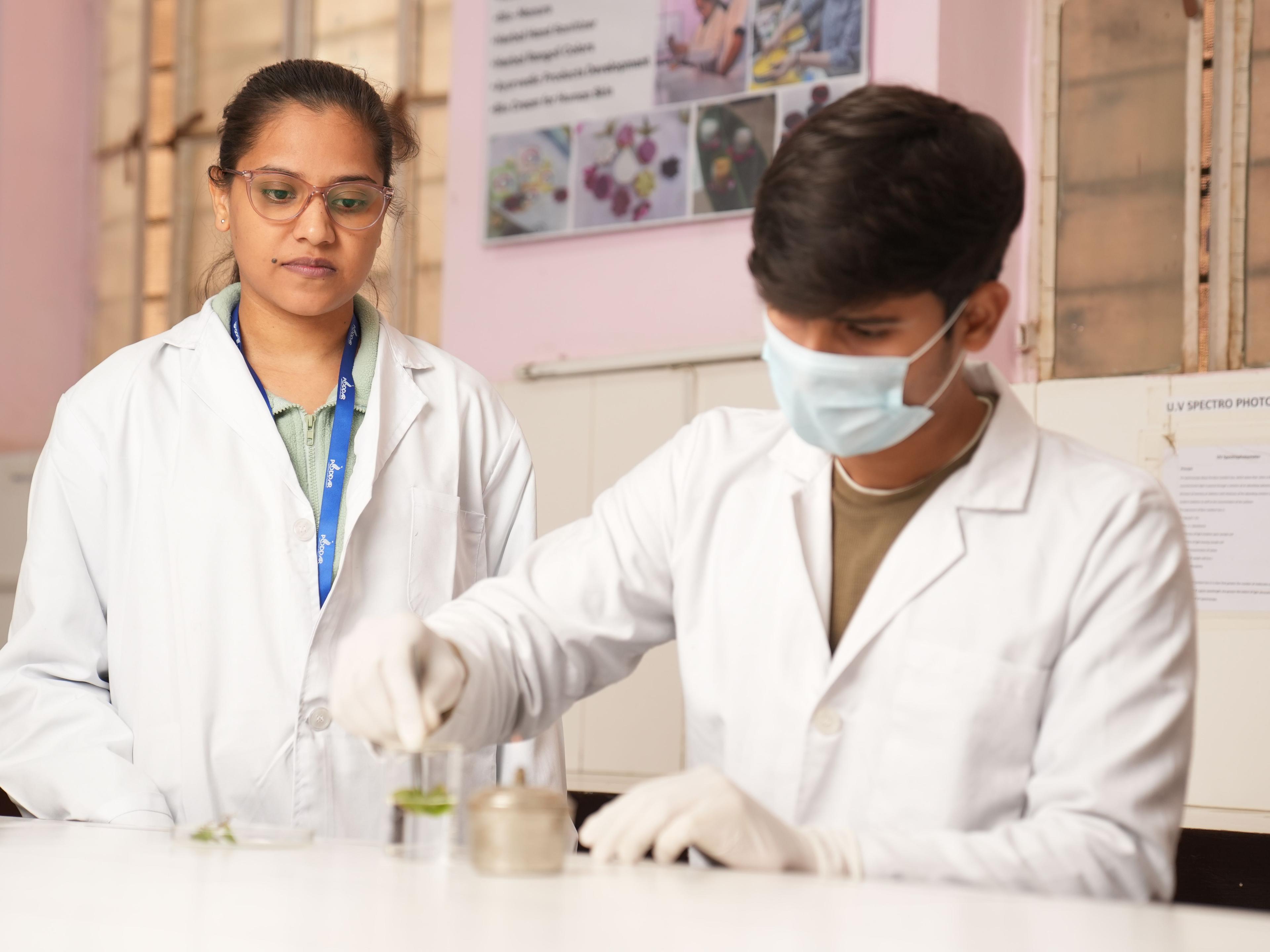 Two scientists in white lab coats and safety gloves working together at a laboratory bench with glassware and research equipment.