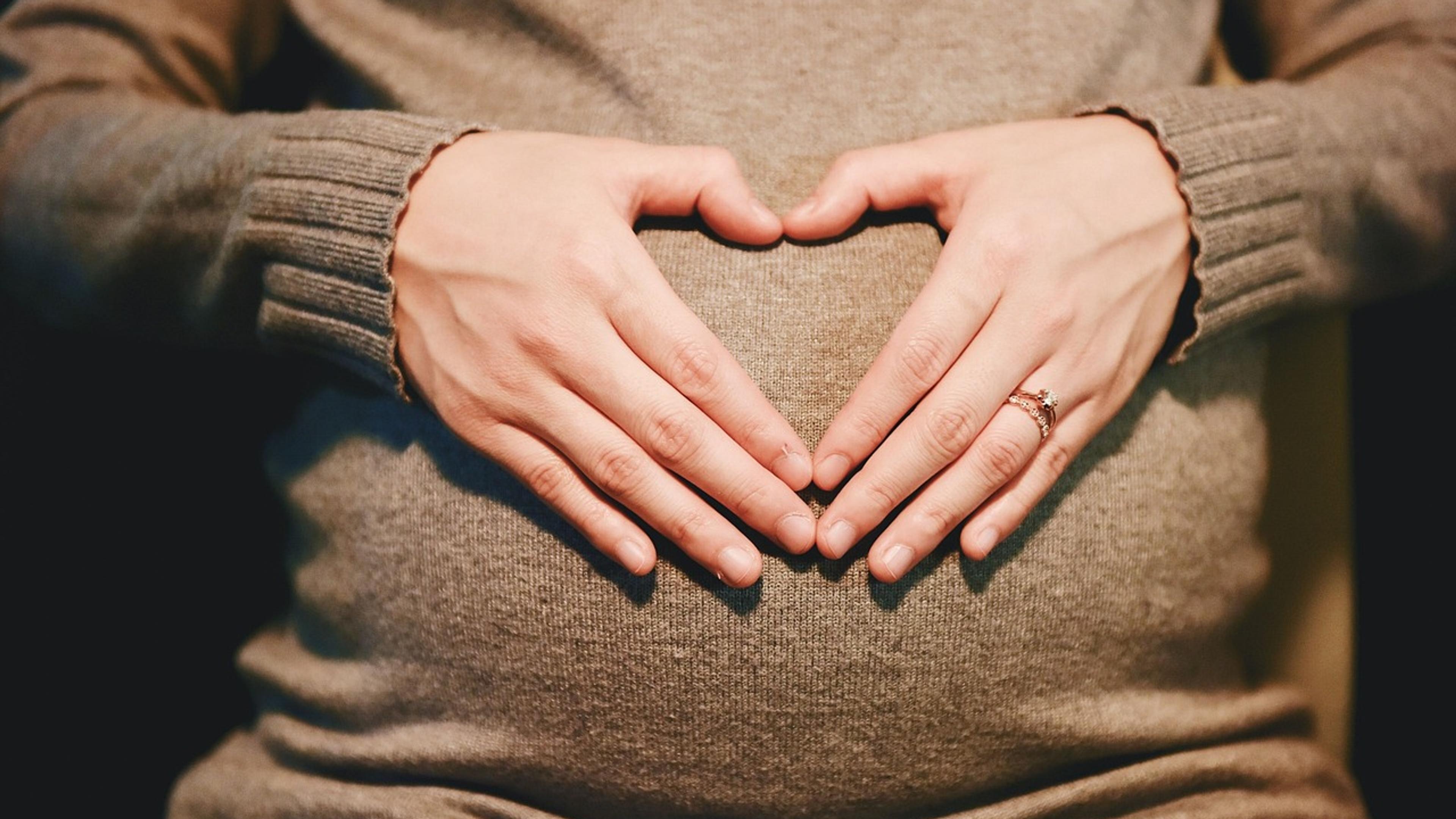 A pregnant woman in a knitted sweater forming a heart shape with her hands over her belly