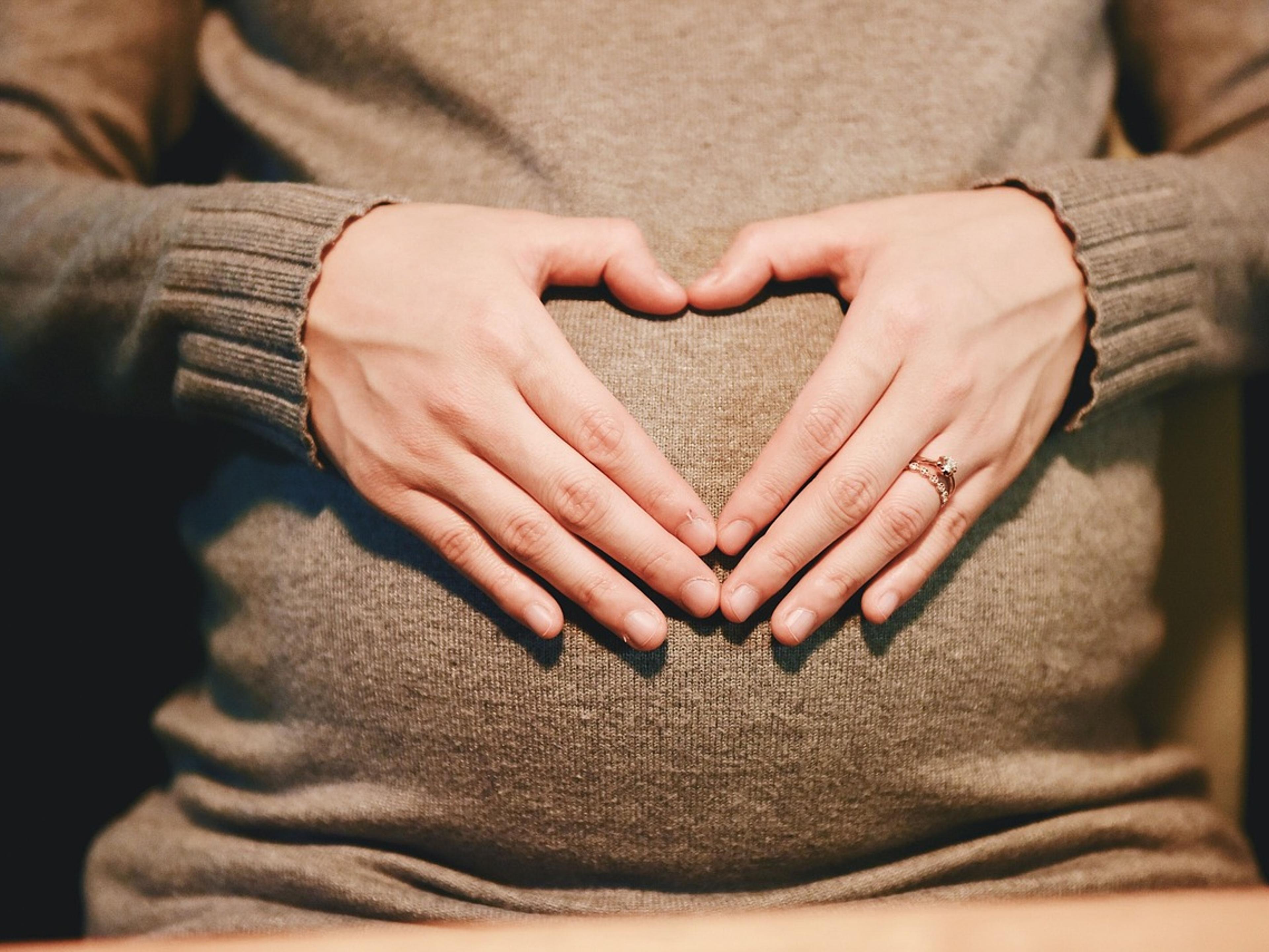 A pregnant woman in a knitted sweater forming a heart shape with her hands over her belly