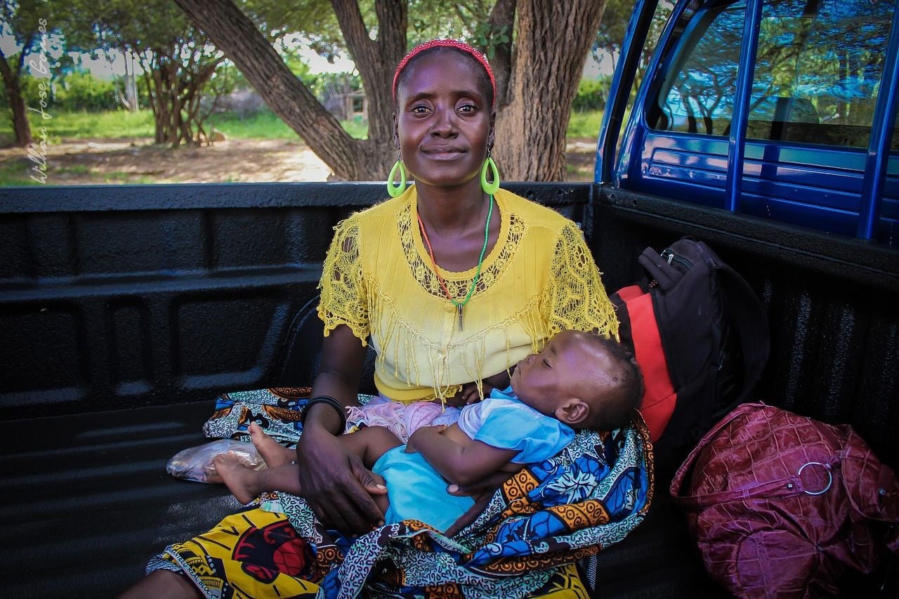 A smiling African woman in a yellow top holding a sleeping baby on her lap