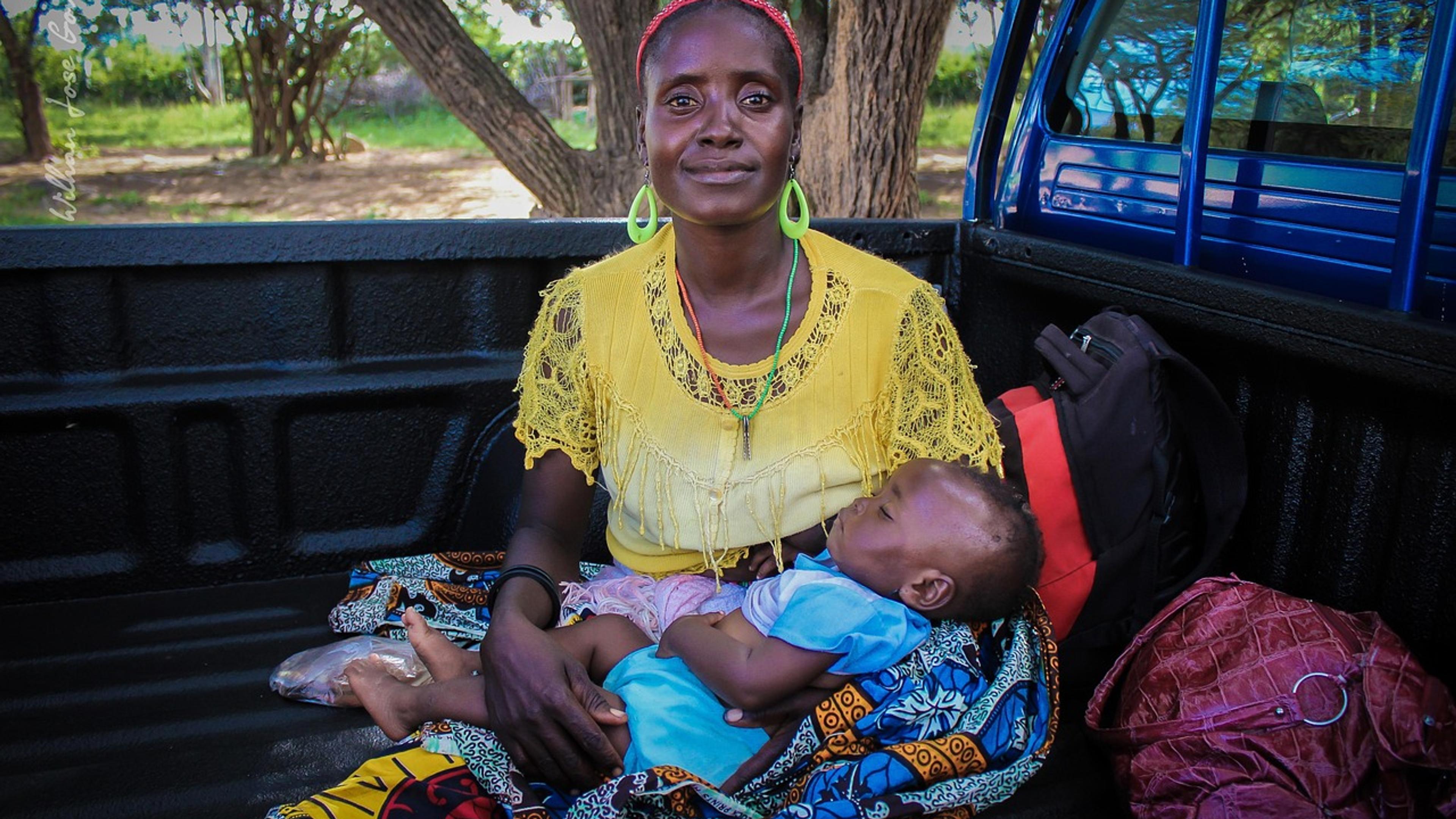A smiling African woman in a yellow top holding a sleeping baby on her lap