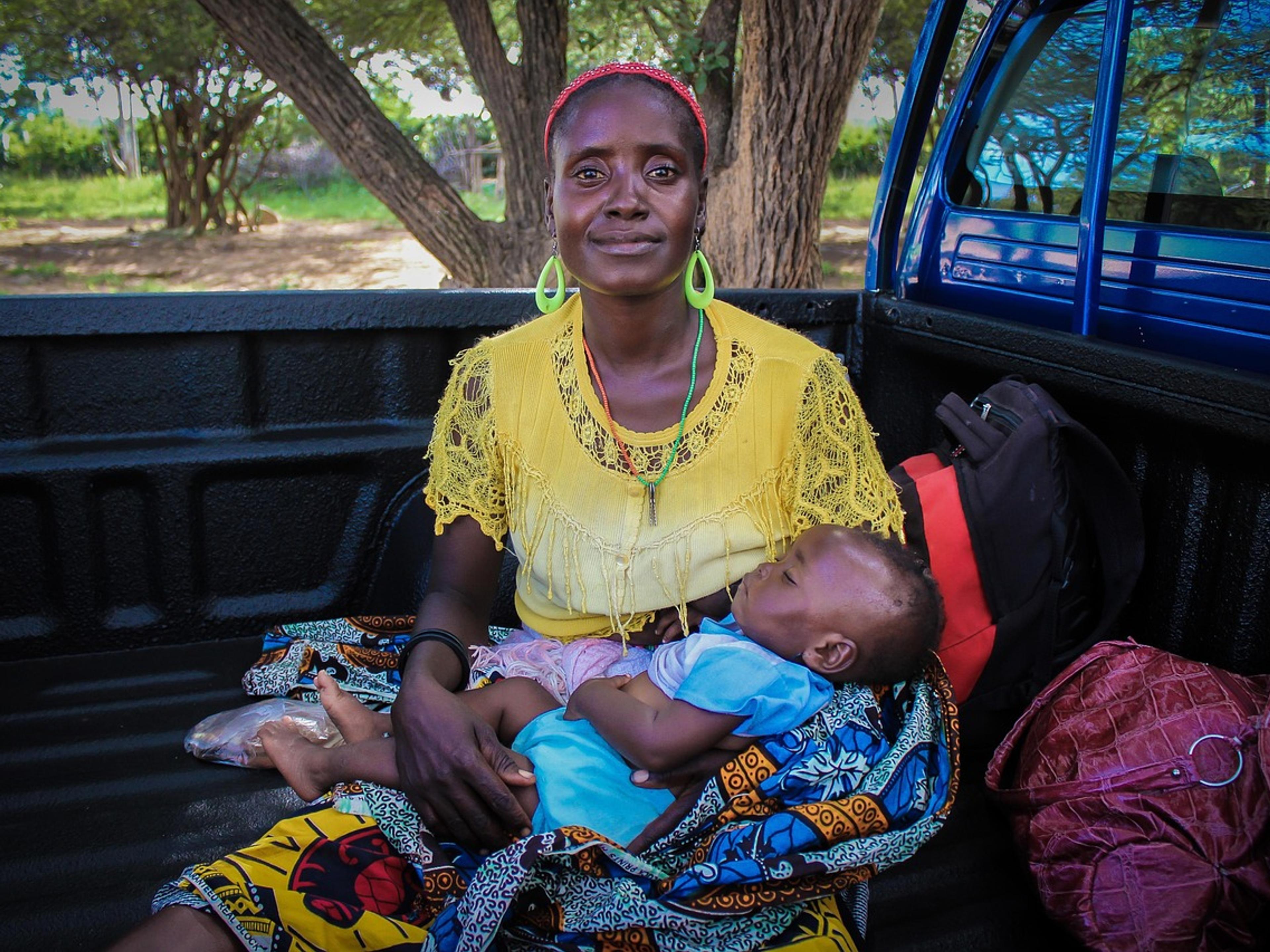 A smiling African woman in a yellow top holding a sleeping baby on her lap