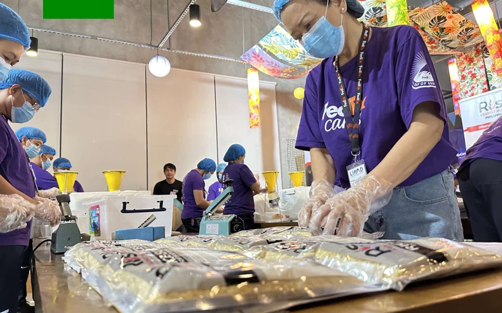 FedEx volunteers in purple shirts, hairnets, face masks and plastic gloves sealing bags of meal packs at a long table